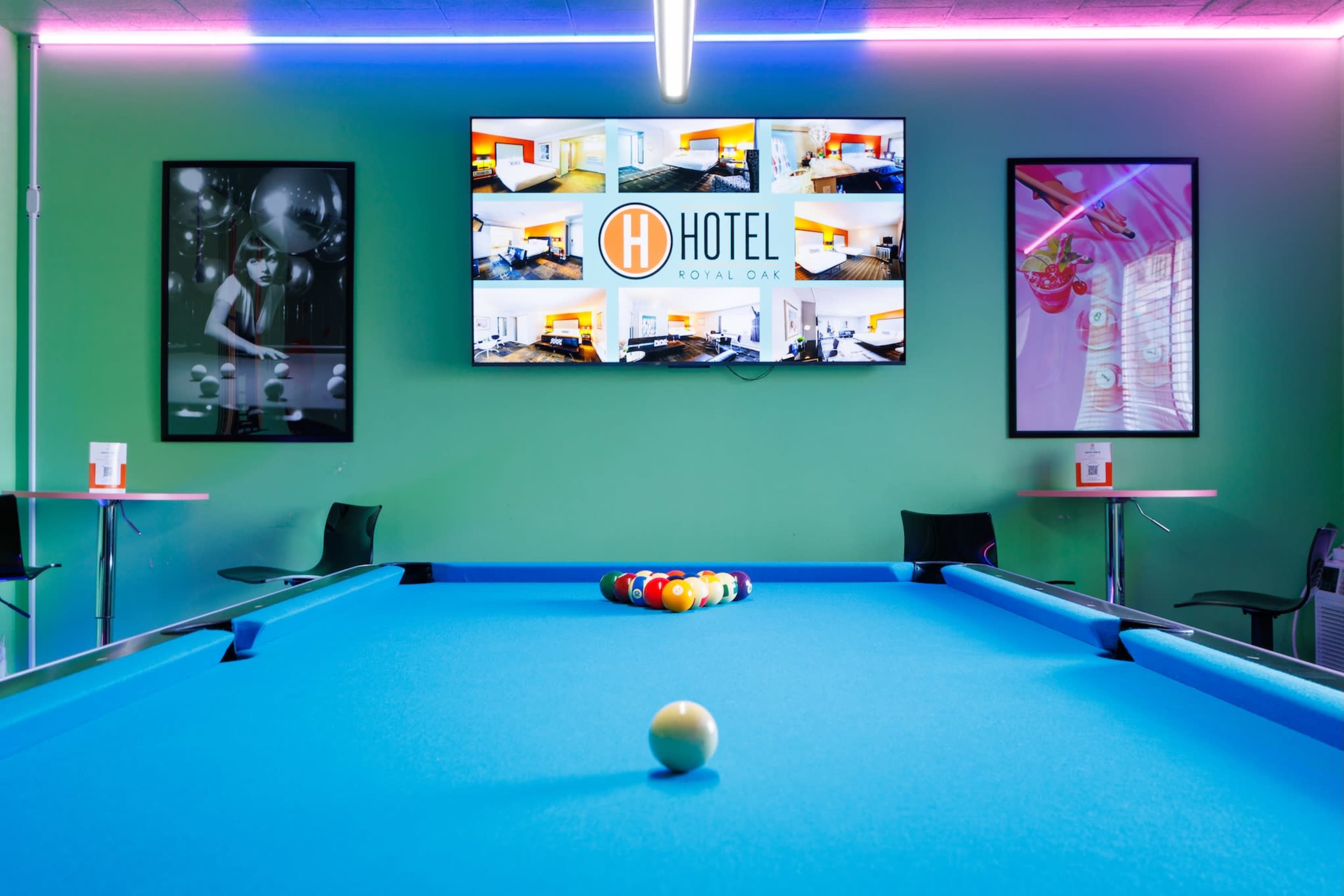 A pool table with colorful balls arranged in a triangular formation is positioned in front of a large screen displaying hotel information, with vibrant green walls and framed photographs on either side.