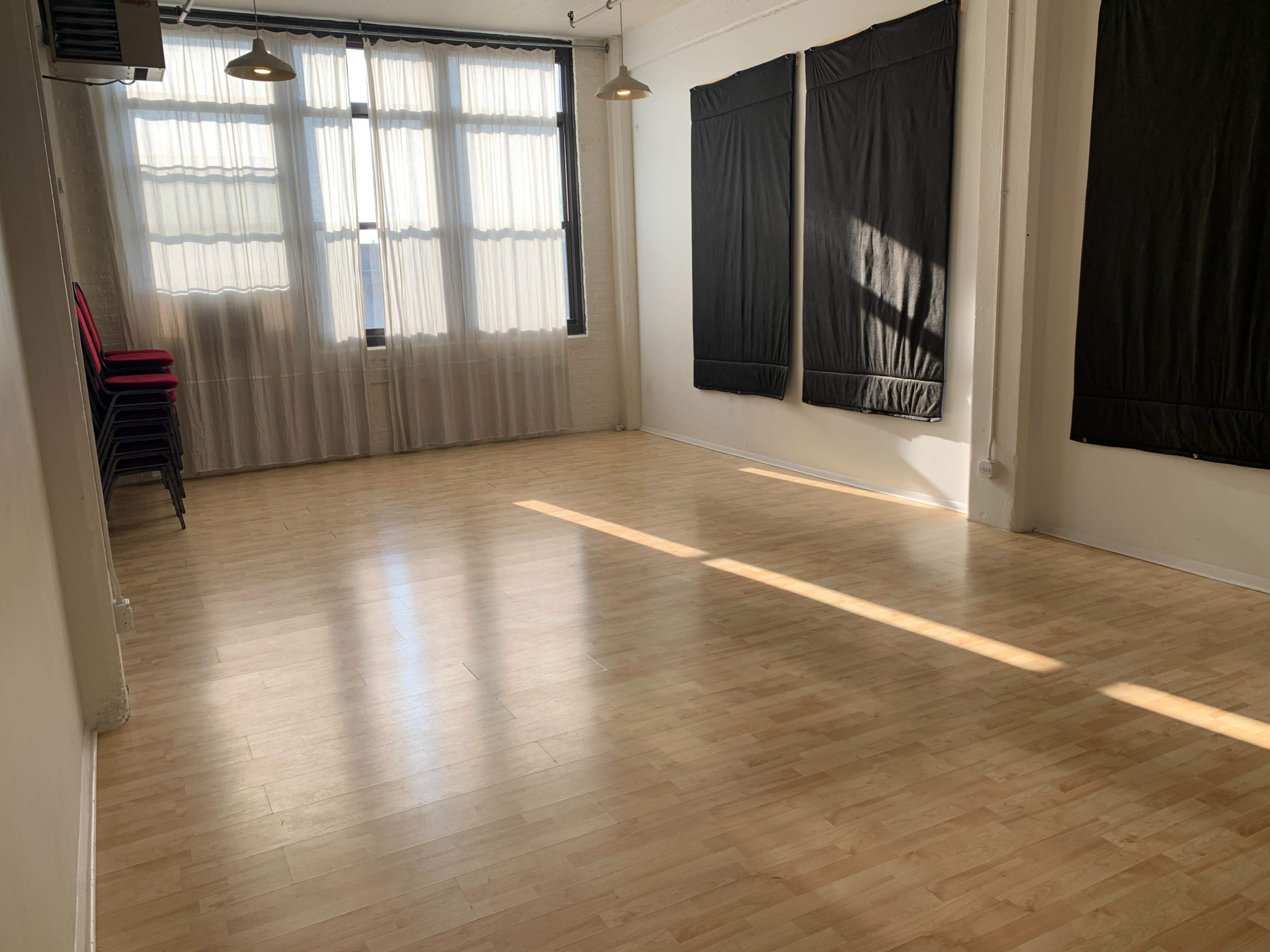 The image shows a spacious, sunlit room with wooden flooring and black fabric-covered walls, featuring stacked chairs in one corner.