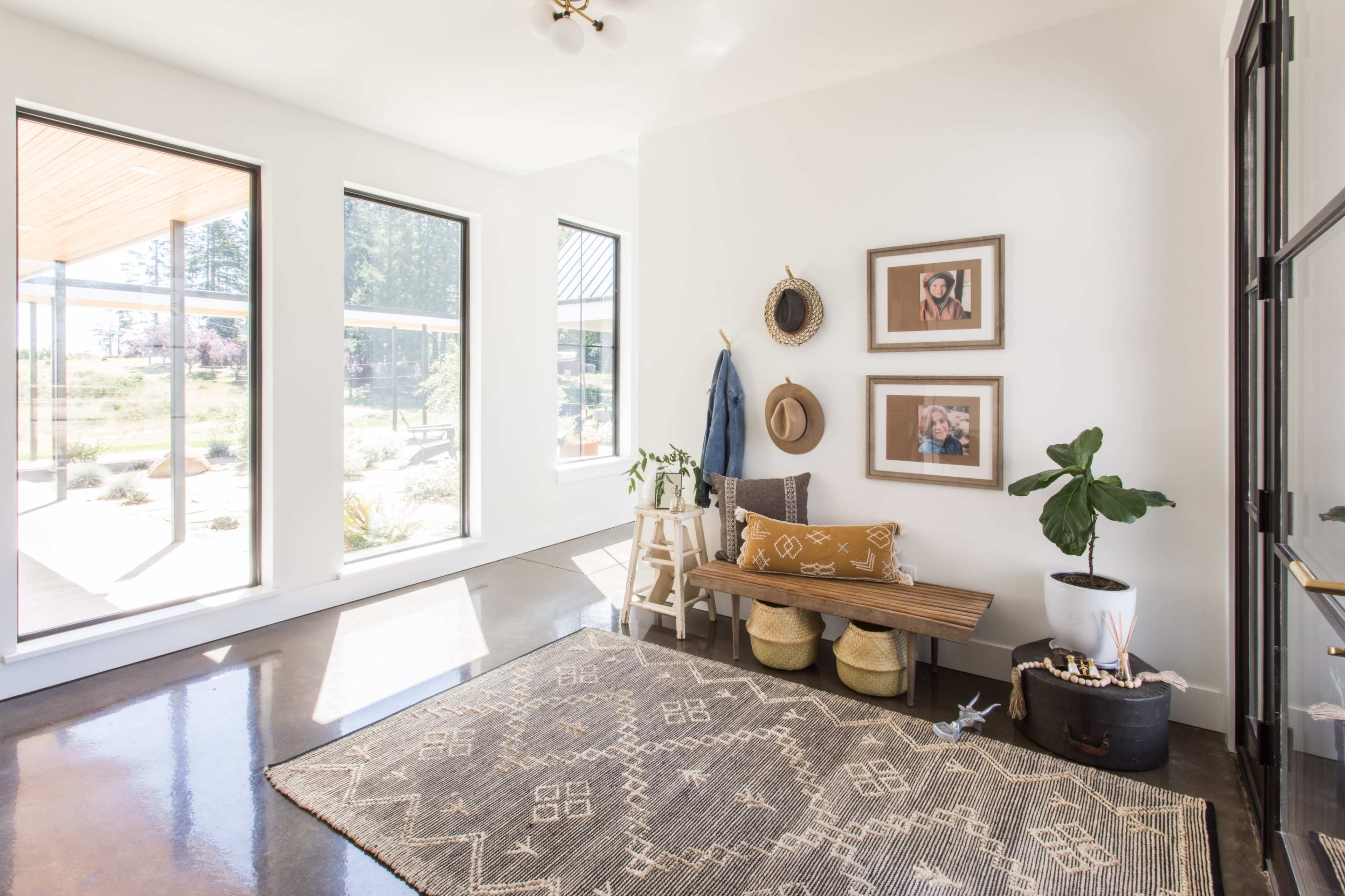A bright entryway with large windows, a wooden bench, framed photos on the wall, a plant, and a patterned rug.