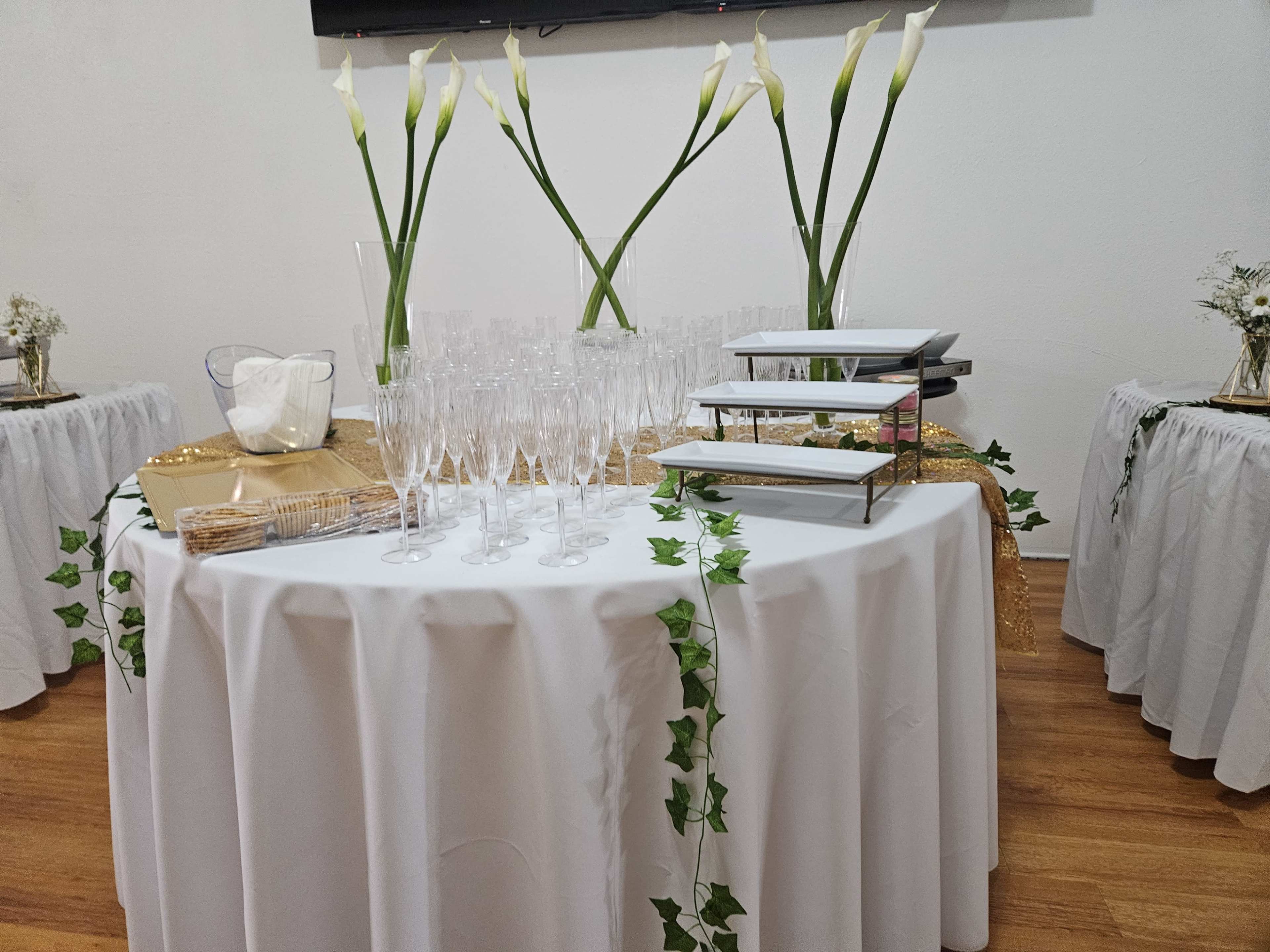 A round table covered with a tablecloth holds glasses, a tiered serving tray, and decorative elements, surrounded by two smaller tables adorned with white tablecloths and floral arrangements.
