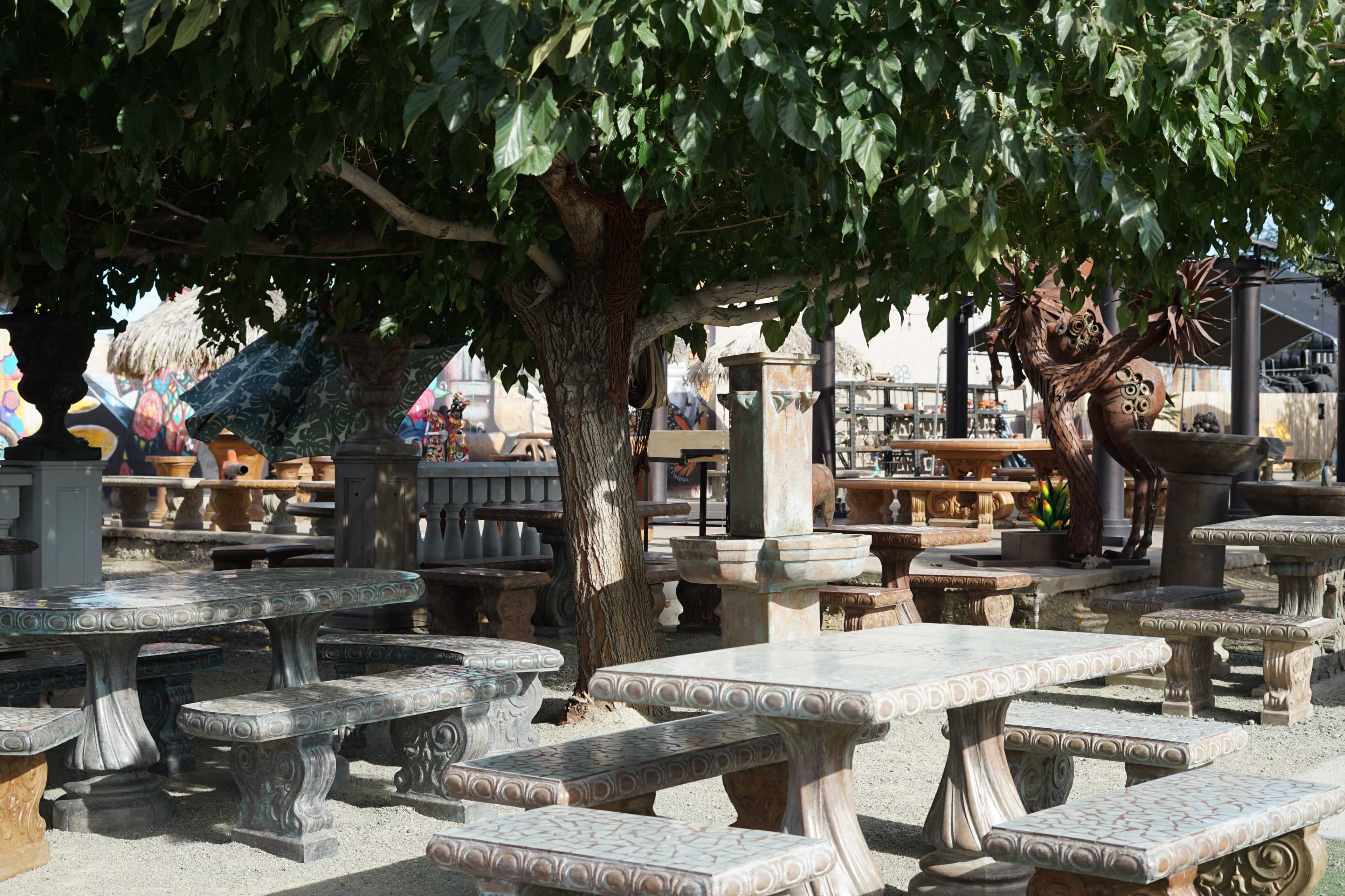 The scene features a collection of stone tables and benches arranged under large trees in an outdoor market.
