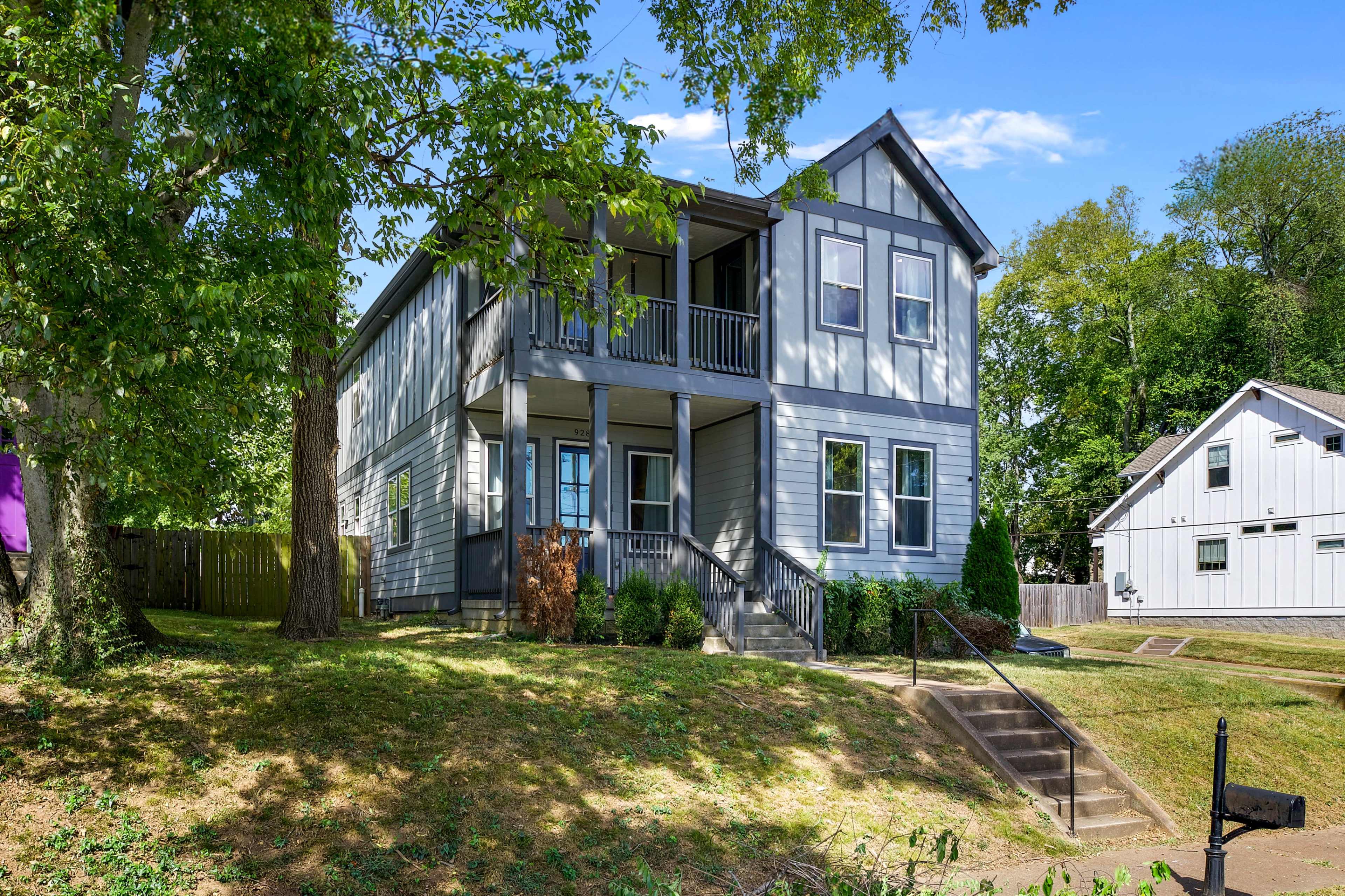 A two-story gray house with a porch and multiple balconies sits among lush greenery in a quiet neighborhood.