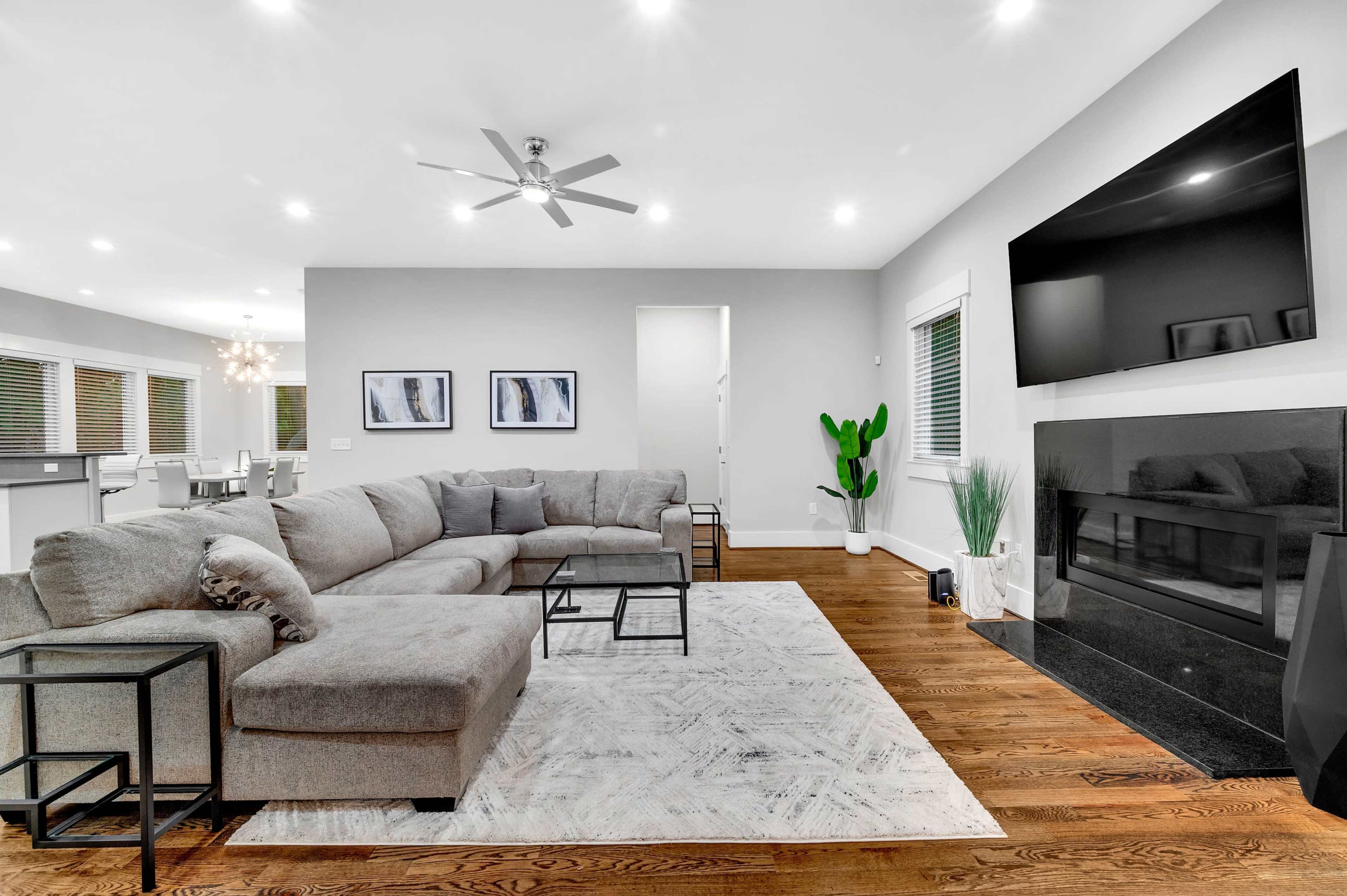 A modern living room featuring a gray sectional sofa, a glass coffee table, a large television mounted on the wall, and a small indoor plant near a fireplace.