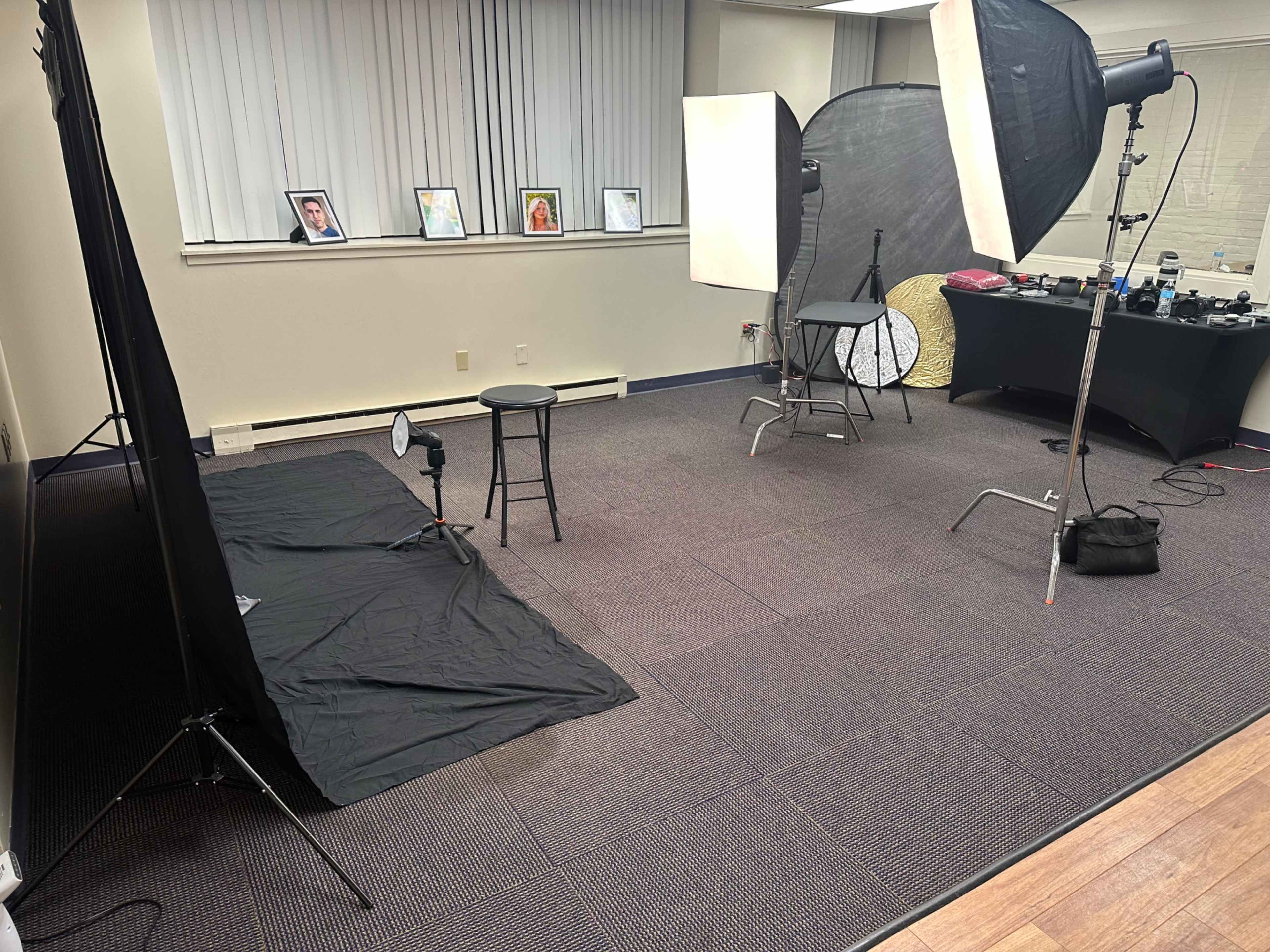 The image shows a photography studio setup featuring two light sources, a black backdrop, a stool, and several framed pictures on the wall.