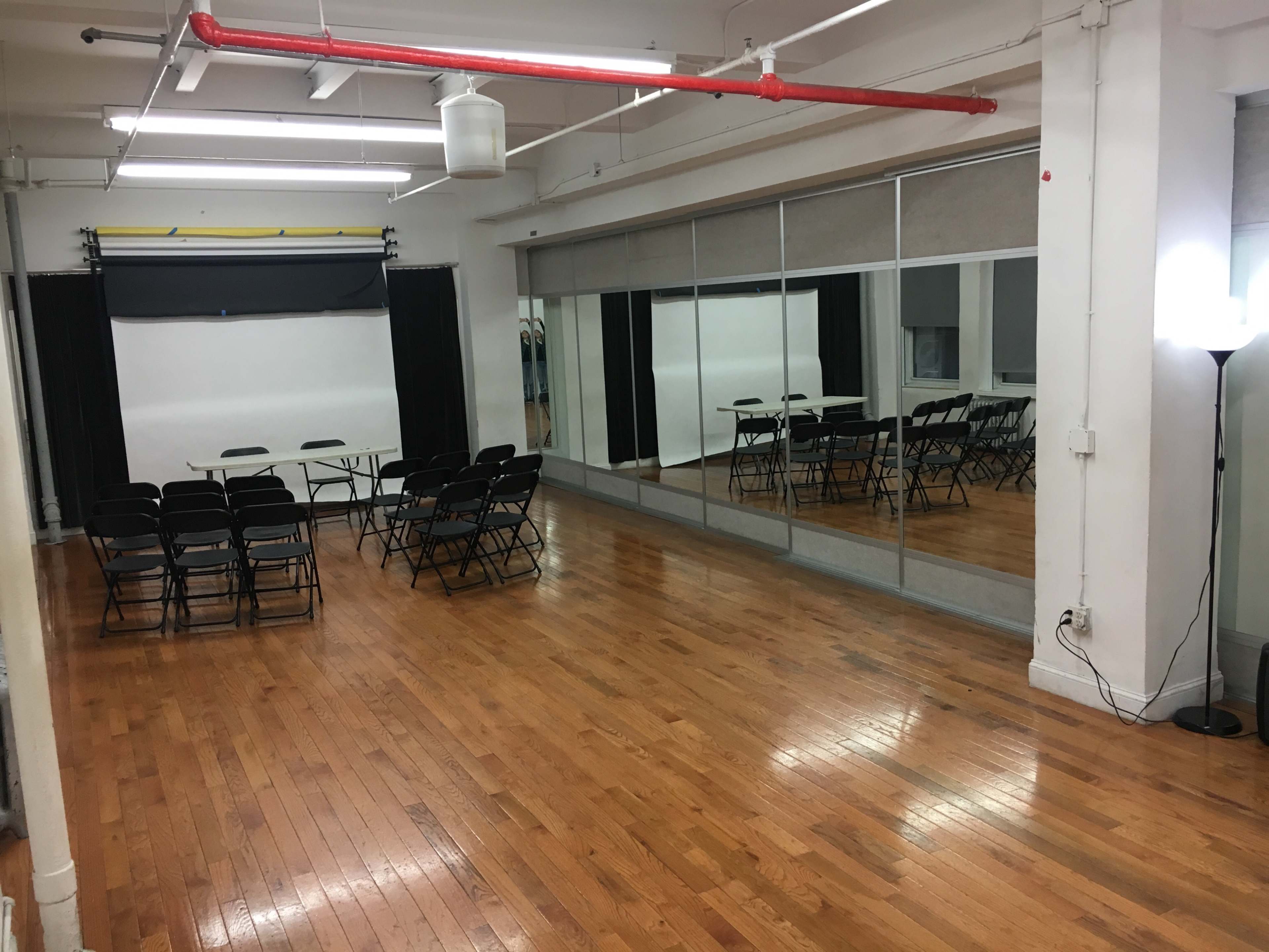 A spacious room features a row of black folding chairs arranged around a table, with large mirrors and a bright light in the corner.