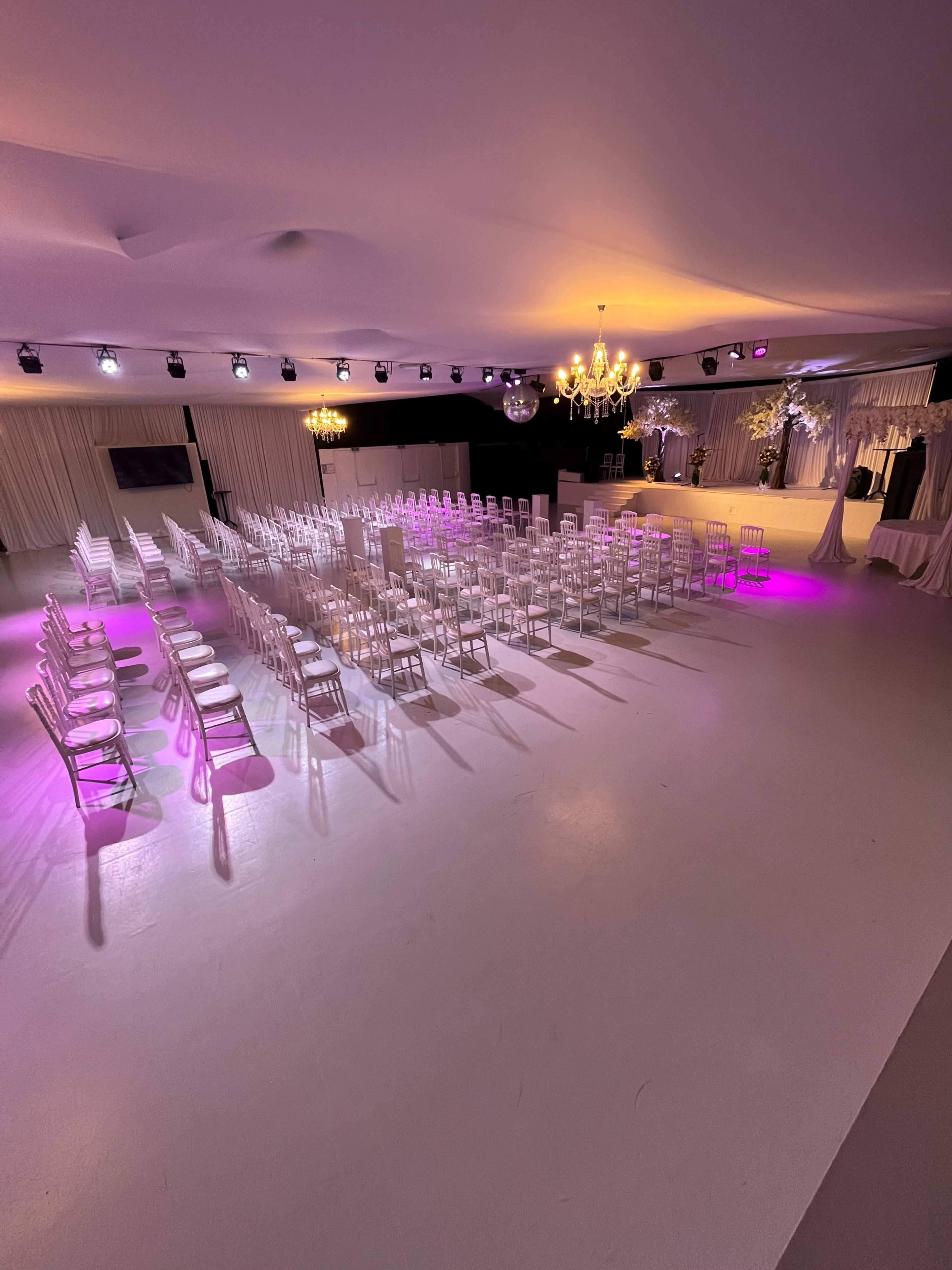 A spacious event venue features rows of white chairs set up for a gathering, illuminated by pink lights and a chandelier overhead.