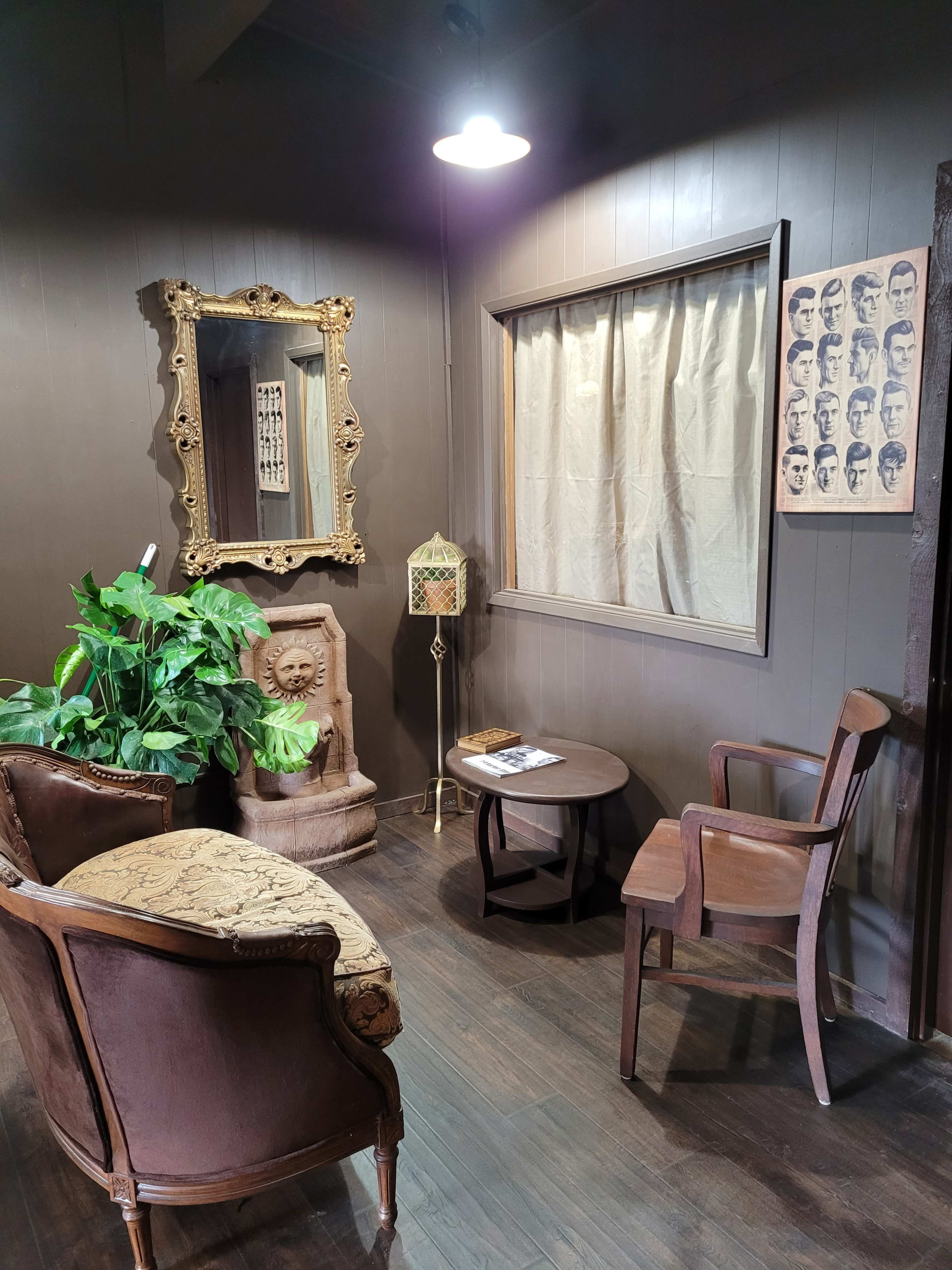 The image shows a cozy corner with a vintage seating arrangement, a mirror, a wall art piece featuring portraits, and a small table surrounded by green plants.