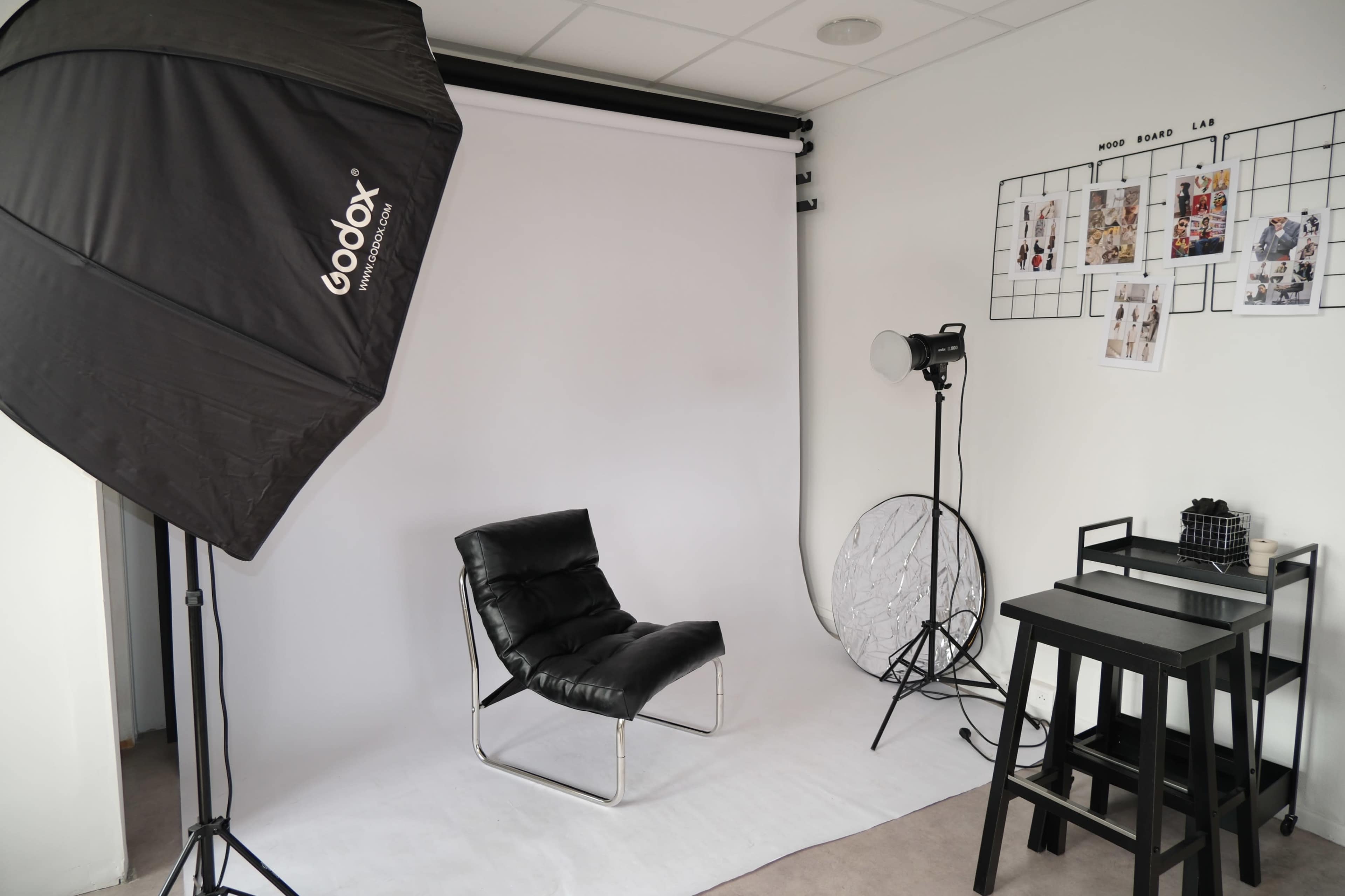 A photography studio setup features a black chair on a white backdrop, with softbox lighting and a few props on a black table.