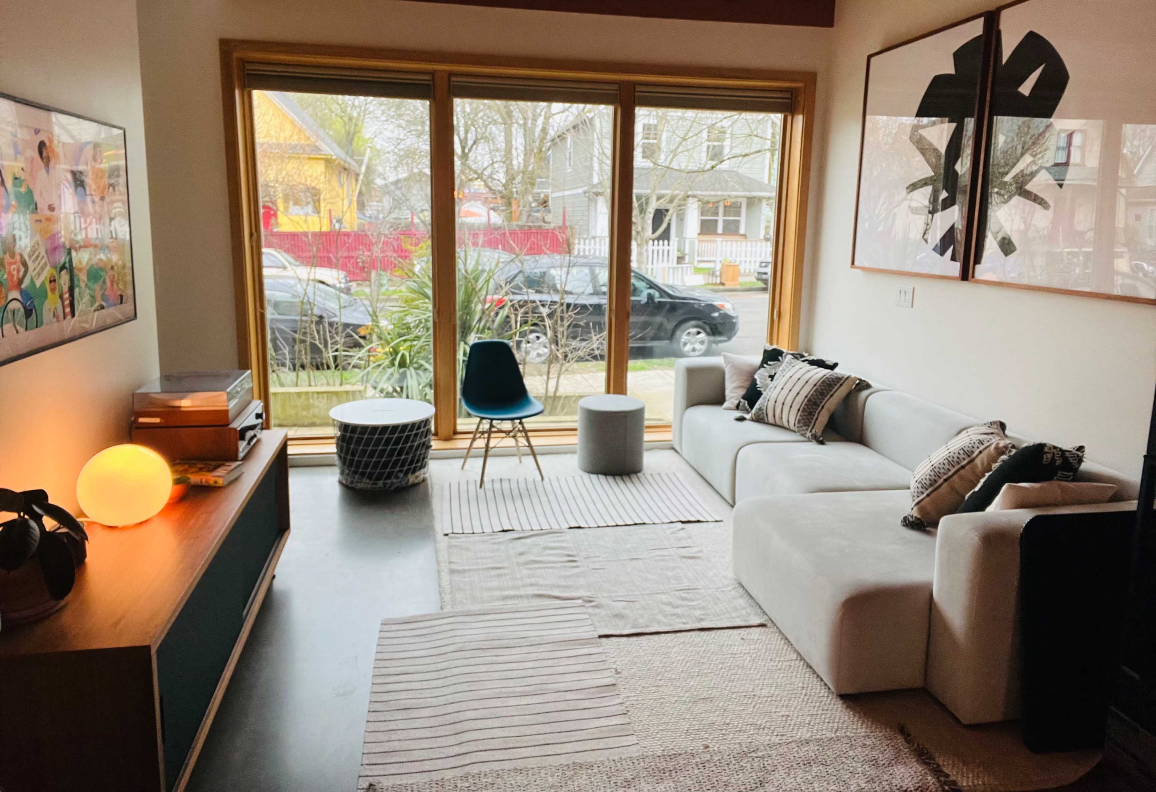 A modern living room features a white sectional sofa, a blue chair, and large windows overlooking a street with parked cars.