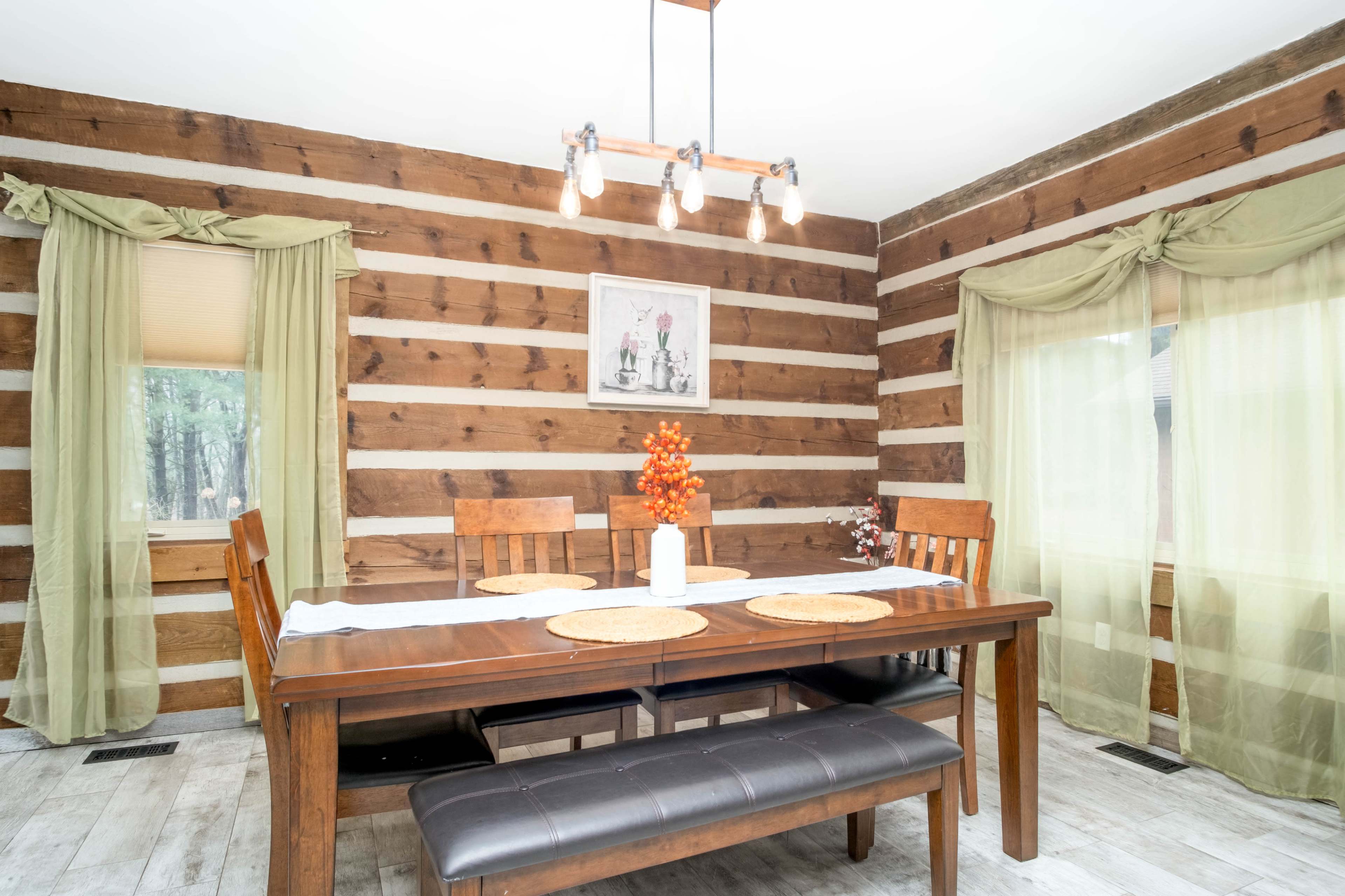 A wooden dining table is set in a cozy room with log cabin walls and large windows adorned with sheer curtains.