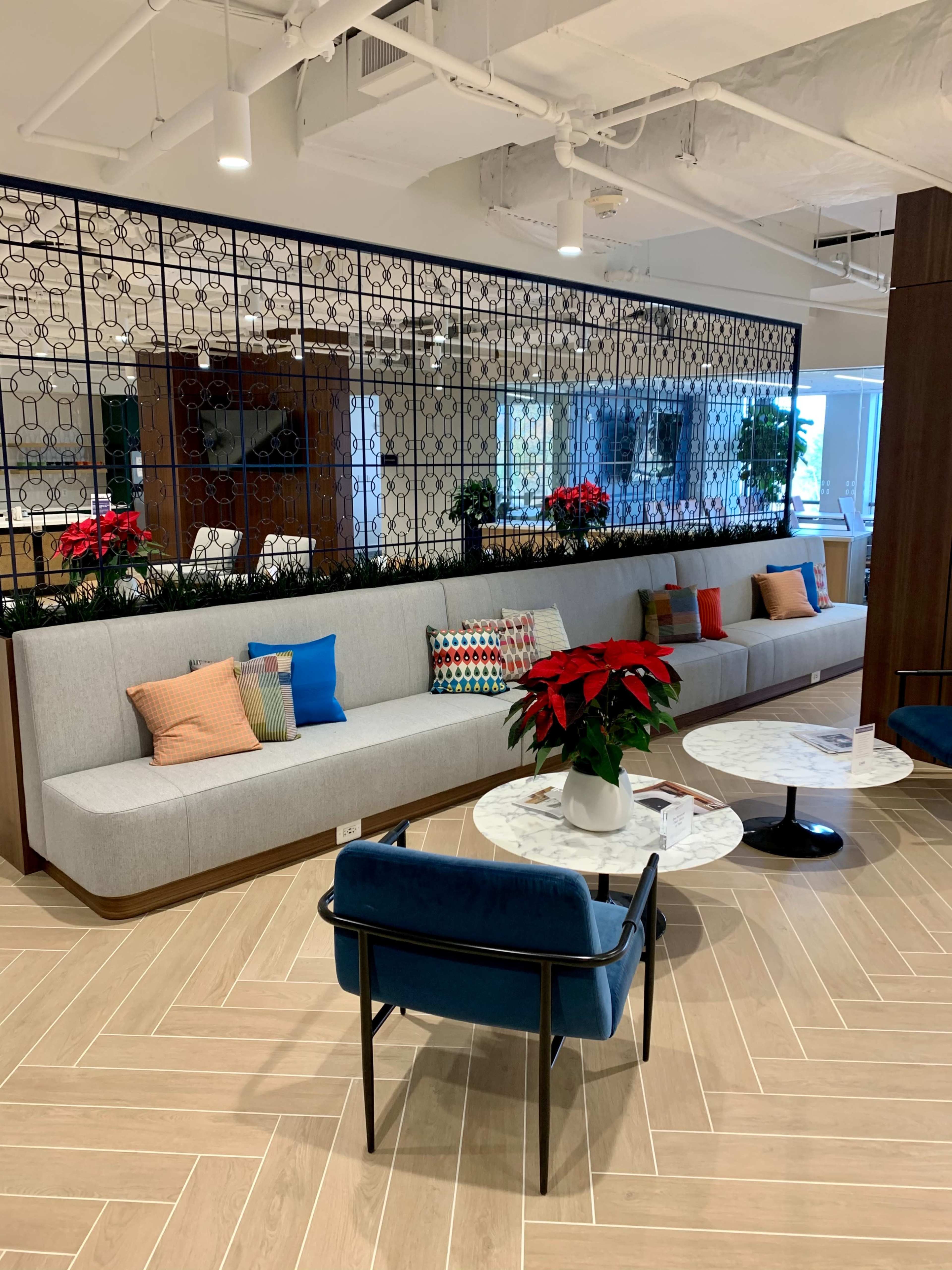 The image shows a modern office lounge area featuring a gray upholstered bench with colorful pillows, a small round table, and a decorative partition with plants and a patterned backdrop.