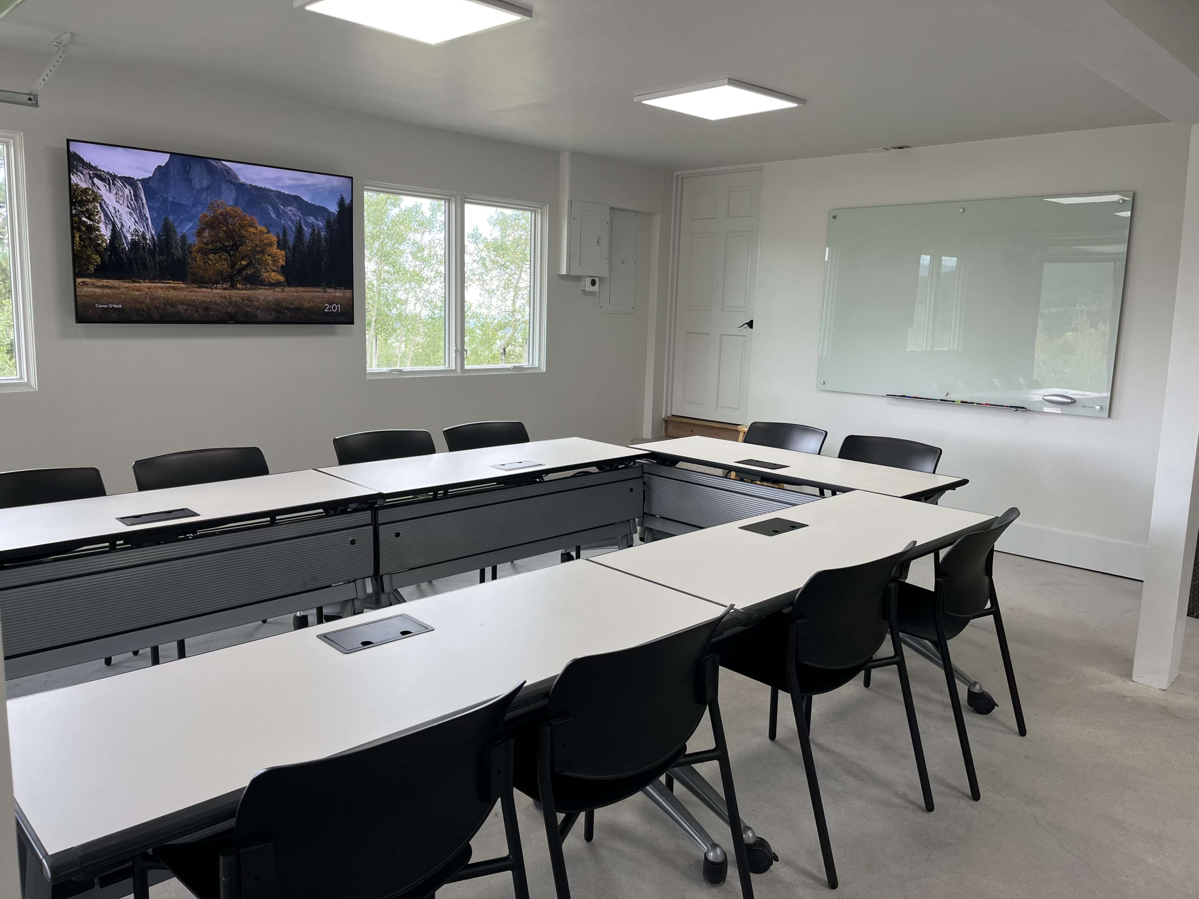 A modern conference room with a large screen, multiple whiteboards, and a setup of tables arranged in an oval shape surrounded by black chairs.