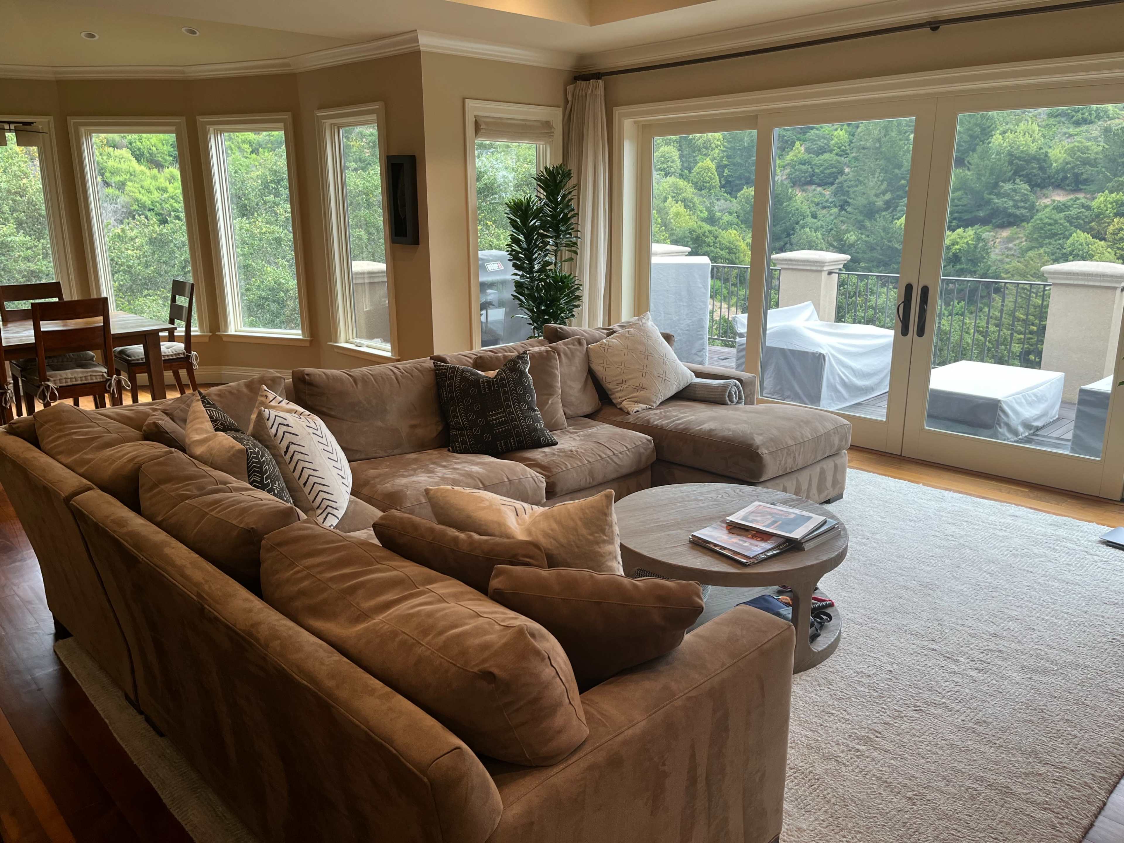 A large, sectional sofa is positioned in a brightly lit living room with windows overlooking a green landscape.