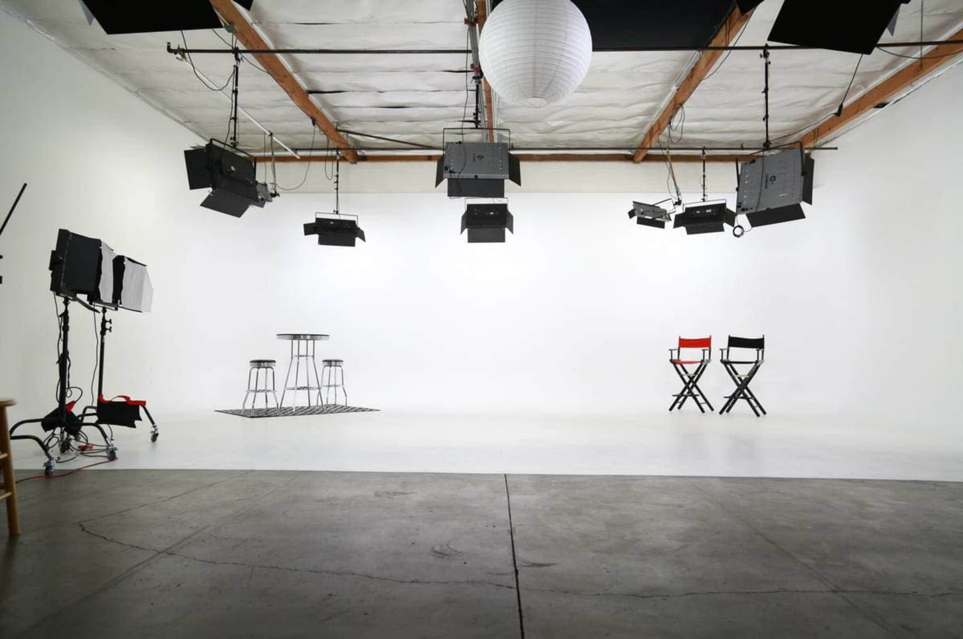 A photography studio features a white backdrop, two chairs, three tall stools, and overhead lighting equipment.