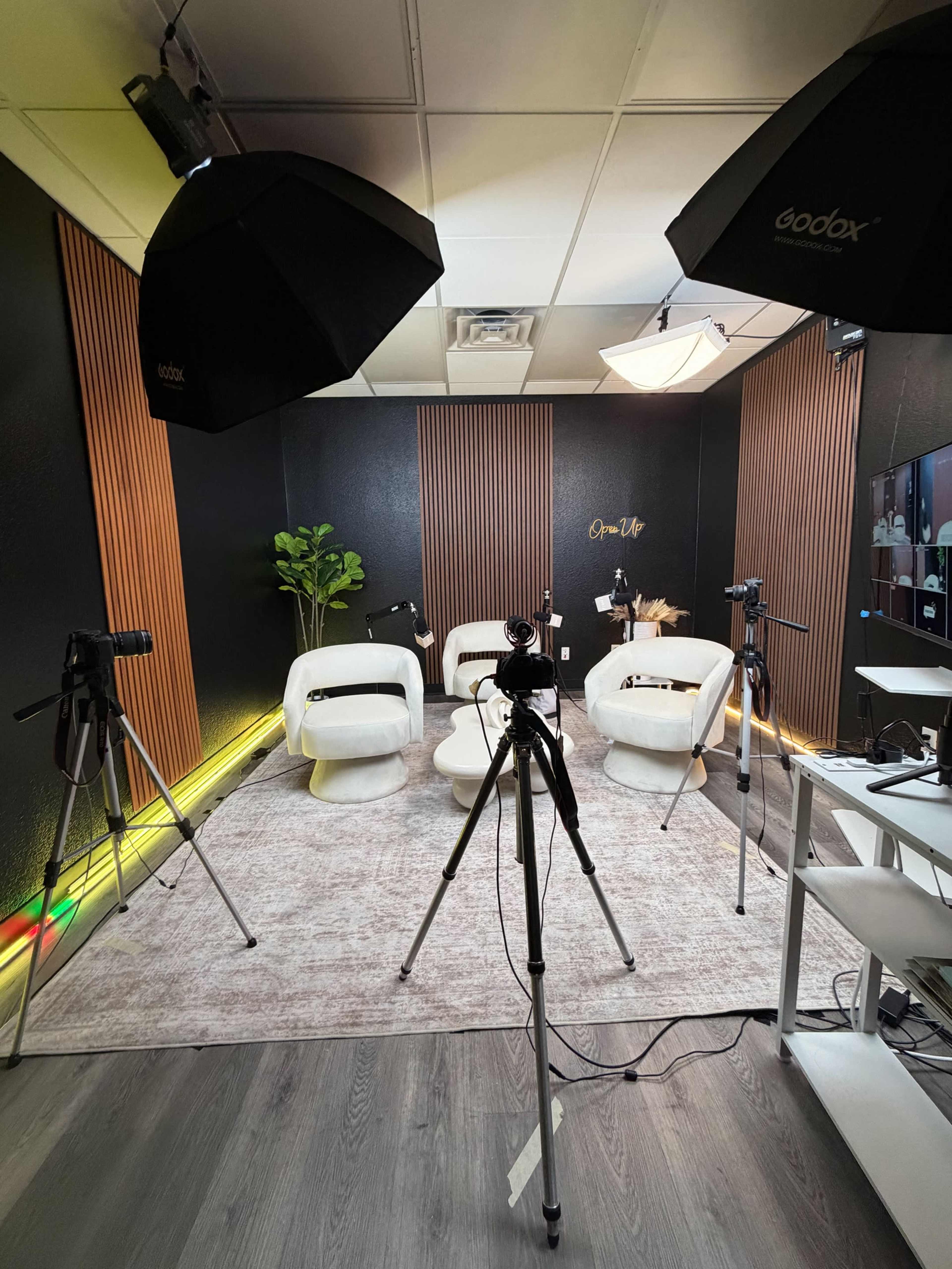 The image shows a modern studio setup with white chairs, soft lighting, and multiple cameras on tripods arranged around a large area rug.