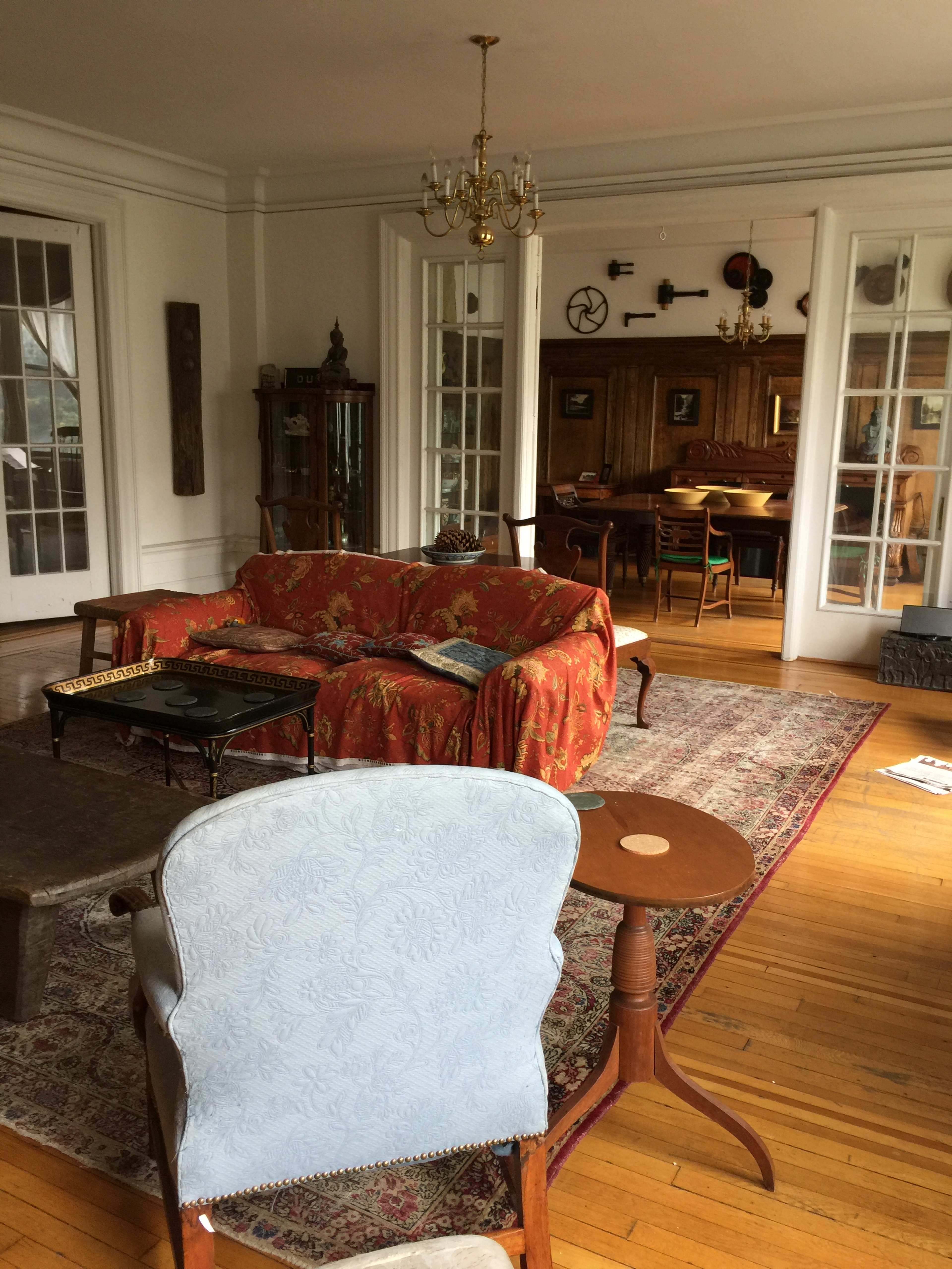 The image shows a spacious, elegantly furnished living room featuring a red floral sofa, a wooden coffee table, and a dining area in the background with several chairs and a wooden sideboard.