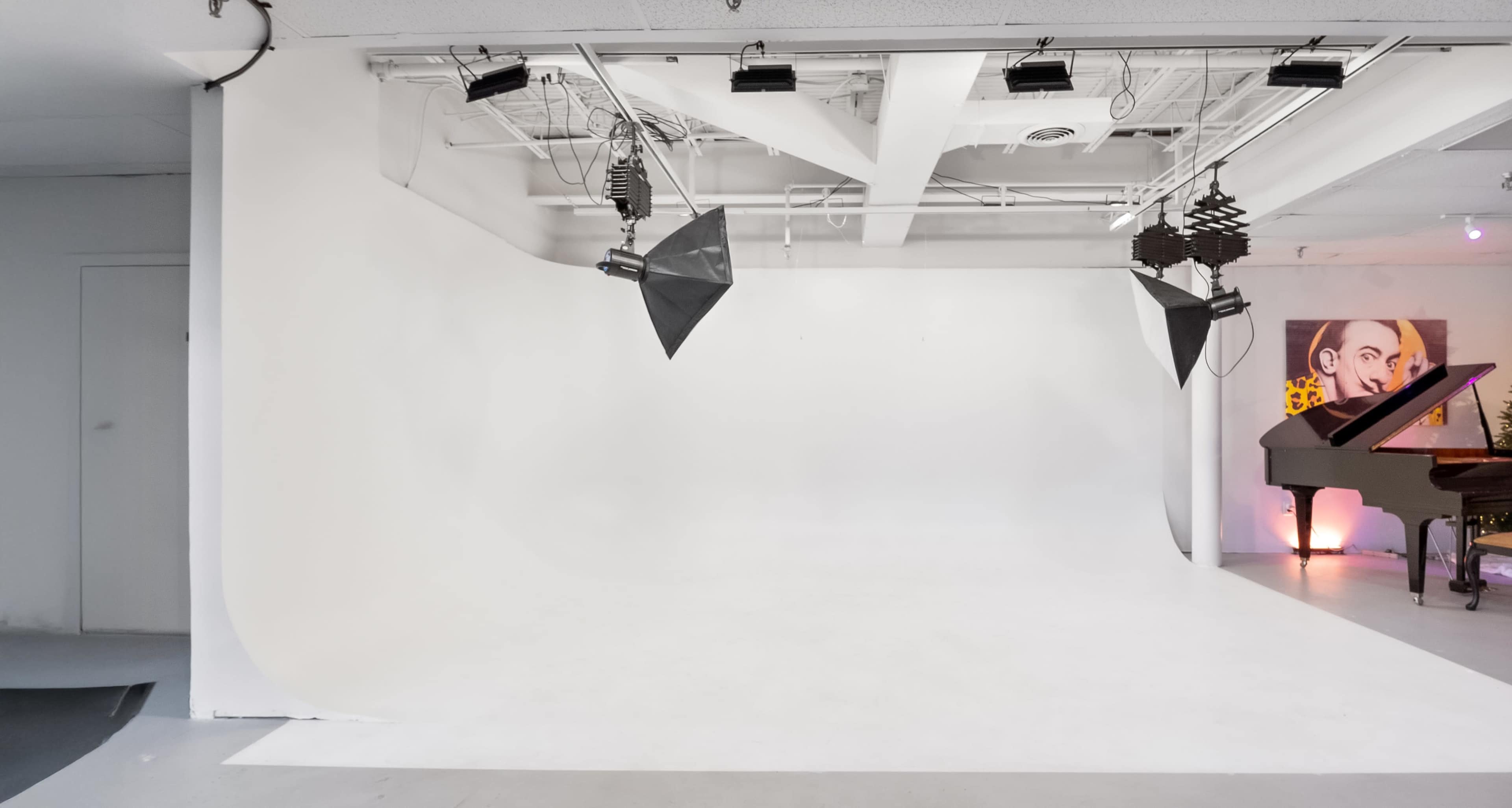 The image shows a spacious photography studio with a seamless white backdrop, studio lights mounted on the ceiling, and a grand piano positioned in one corner.