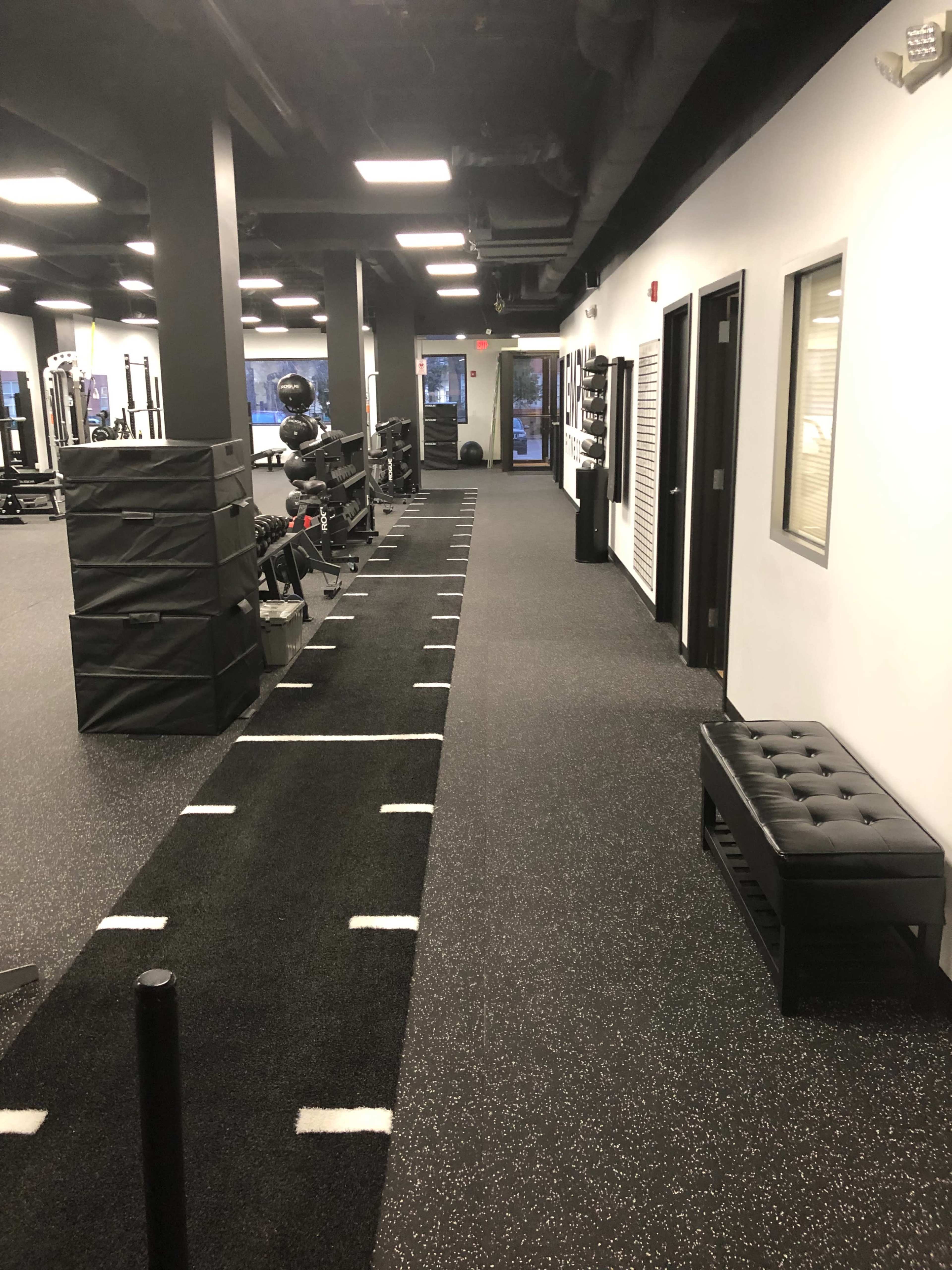 The image shows a well-lit fitness facility with exercise equipment lined against one wall and a long strip of turf running down the middle.