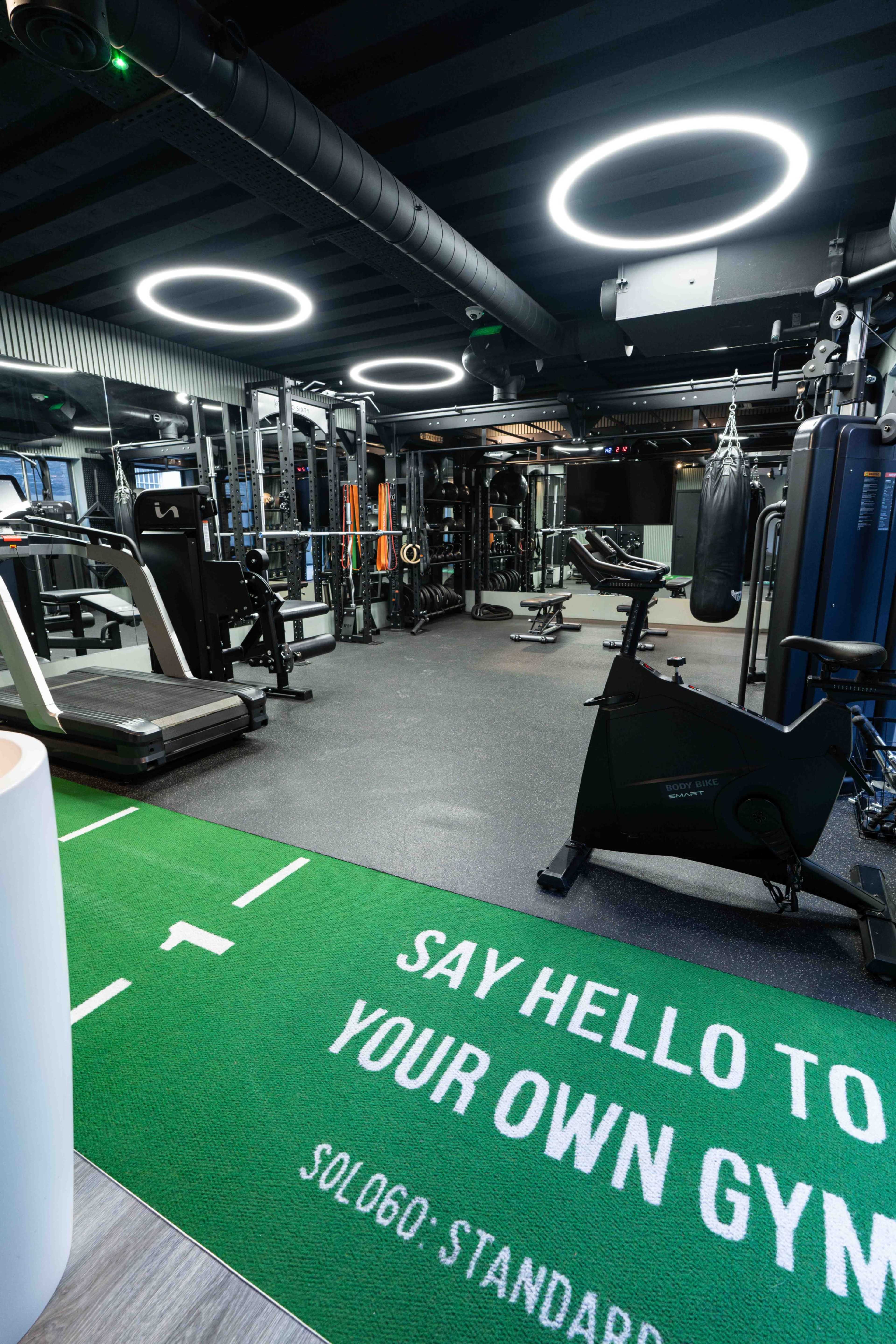 The image shows a modern gym filled with various fitness equipment, including treadmills, weights, and boxing bags, under circular ceiling lights.