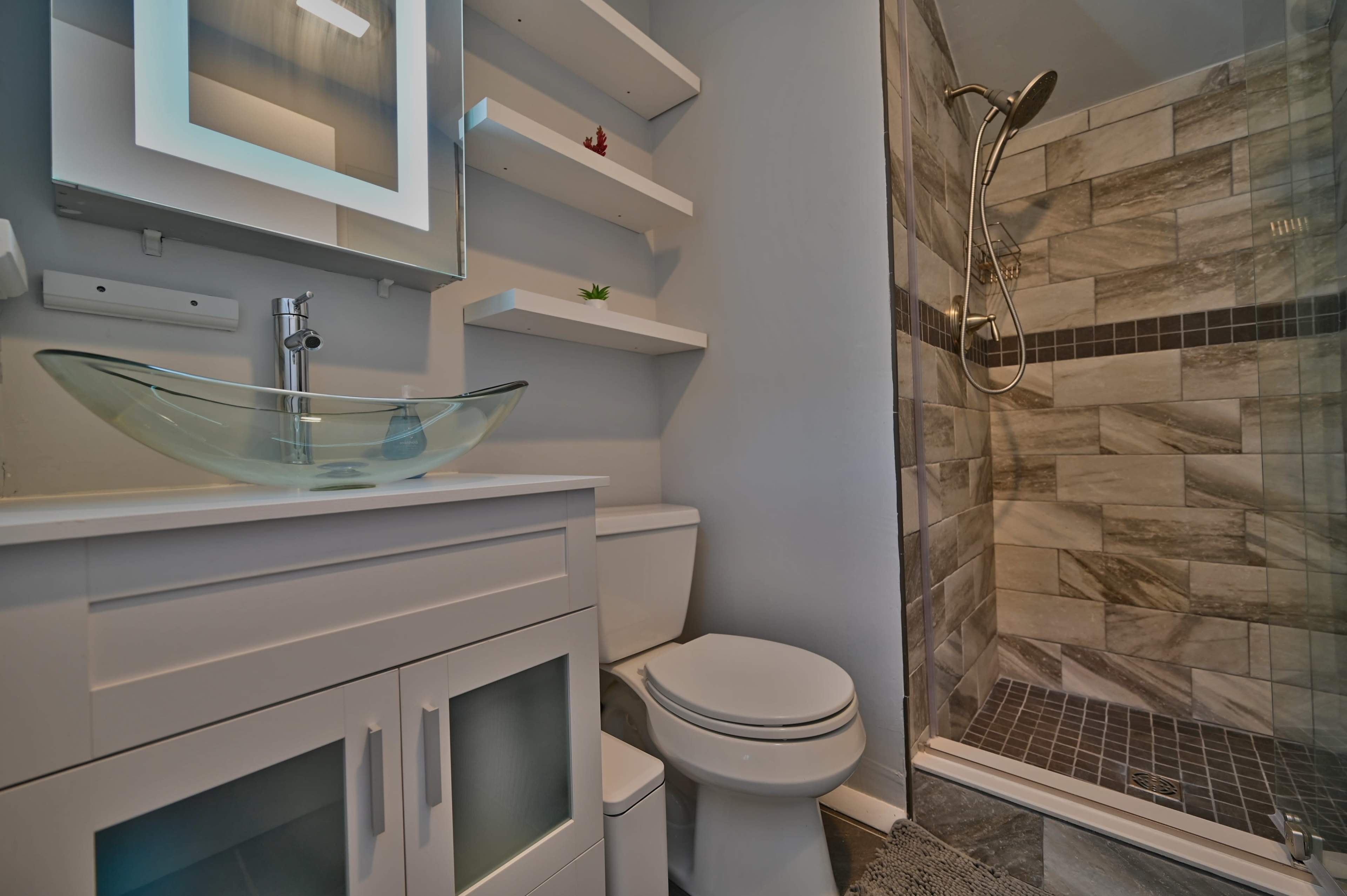 A modern bathroom featuring a glass vessel sink on a white vanity, a toilet, open shelves with decorative items, and a tiled shower area.
