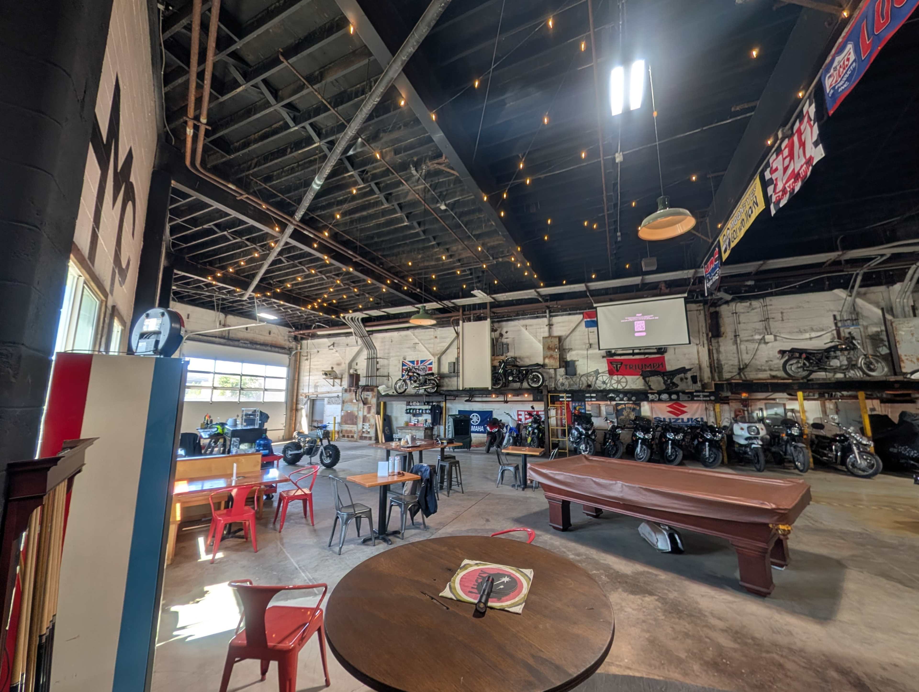 The image depicts a spacious motorcycle shop with a pool table, various tables and chairs, and several motorcycles displayed along the walls under soft lighting.