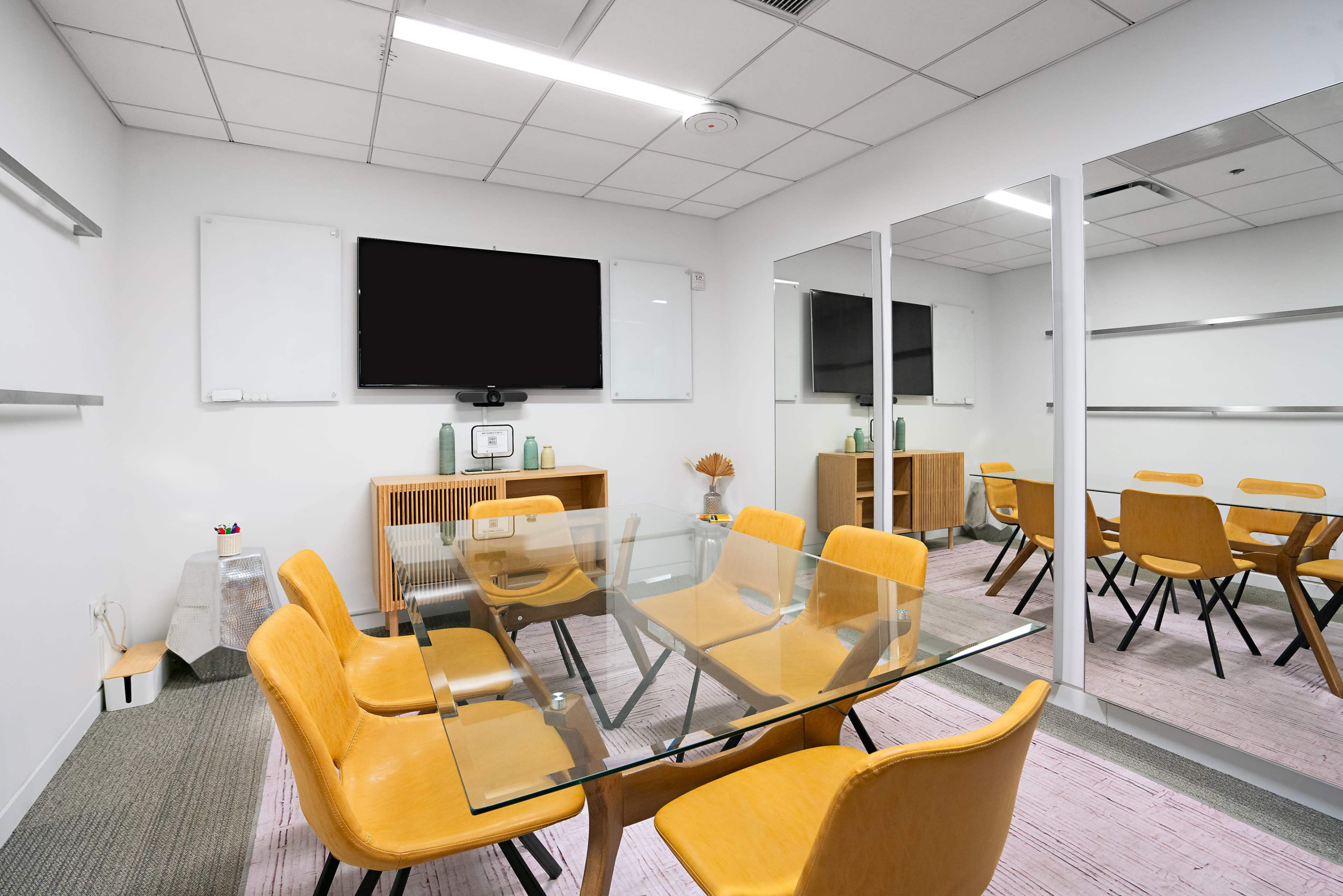 The image shows a modern conference room with a glass table, yellow chairs, a large television screen, and mirrors on one wall.