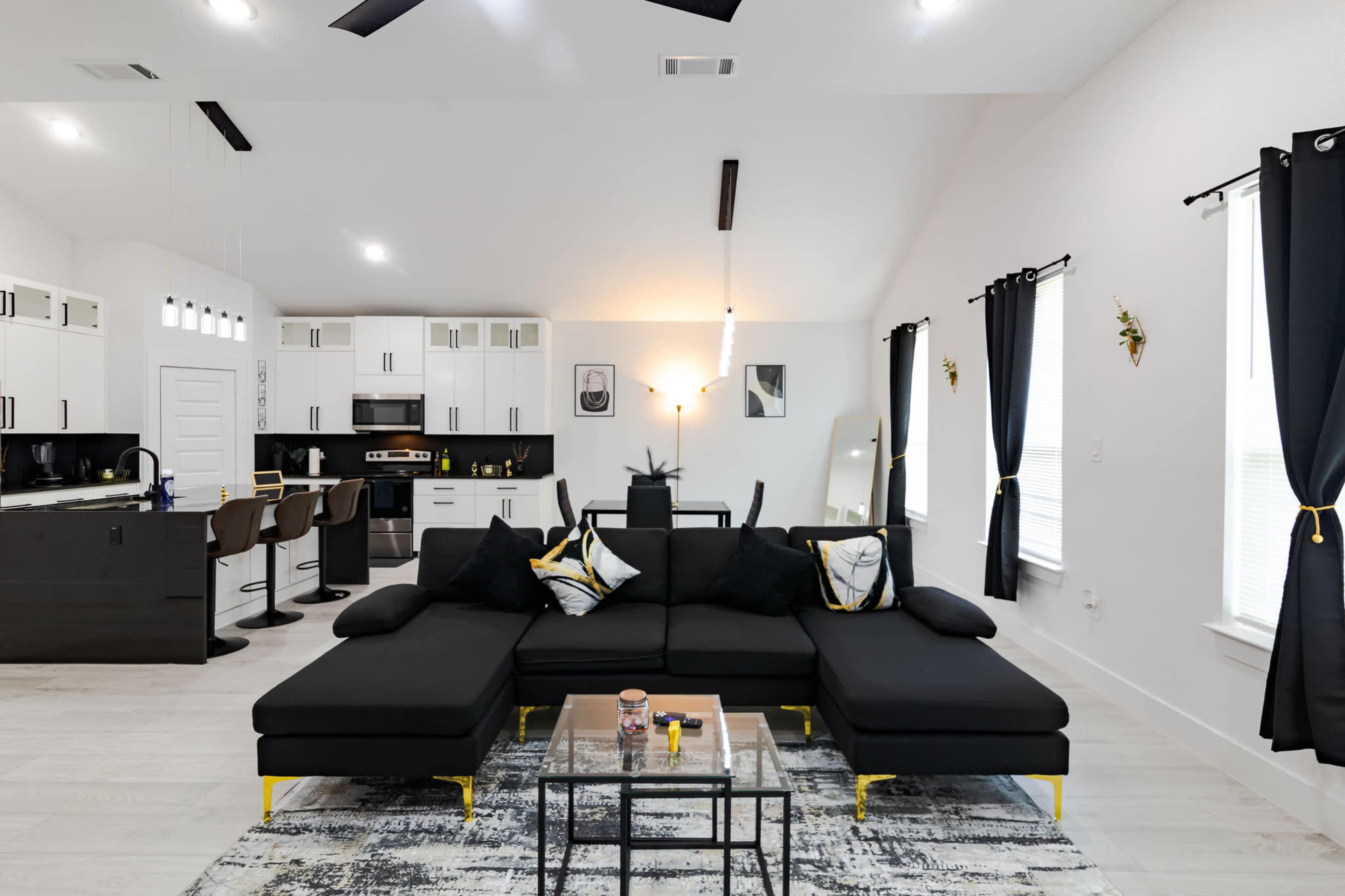 The image shows a modern living room and kitchen area featuring a black sectional sofa, a glass coffee table, and kitchen bar stools against a backdrop of white walls and dark accents.