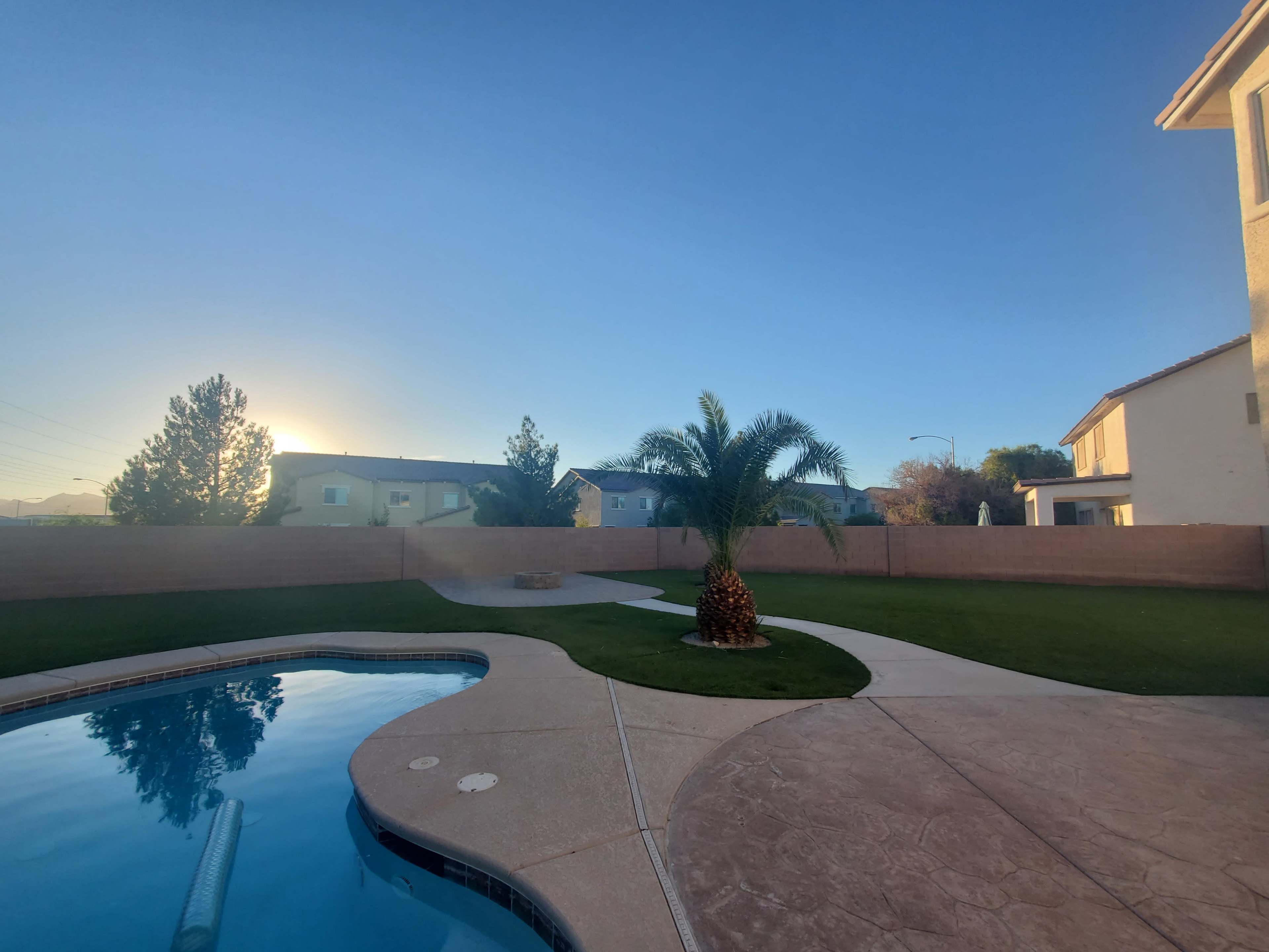 A clear blue sky overlooks a backyard with a swimming pool, palm tree, and well-maintained lawn surrounded by a low wall.