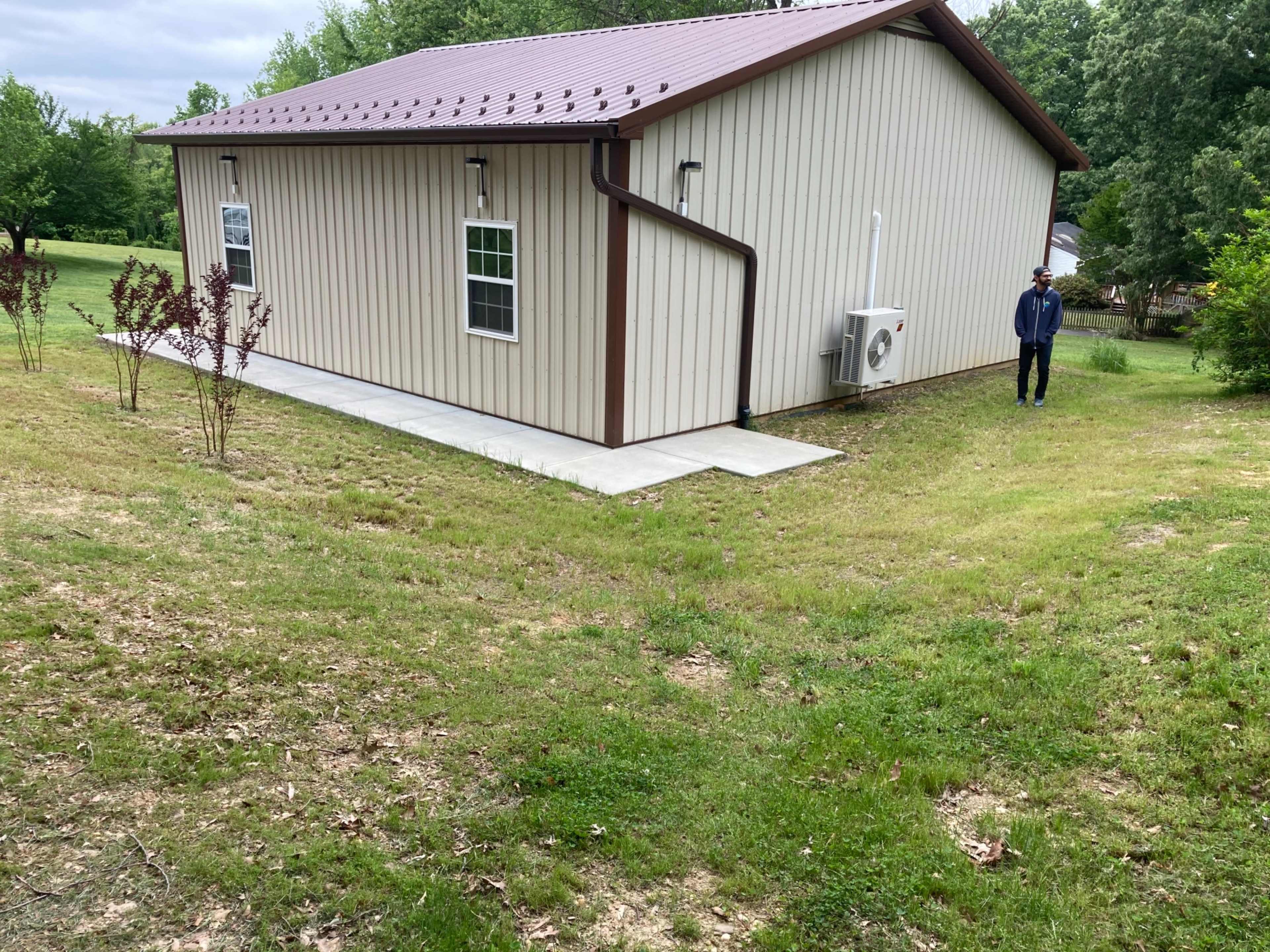 A person stands next to a beige metal building with a sloped lawn leading to a concrete pathway.
