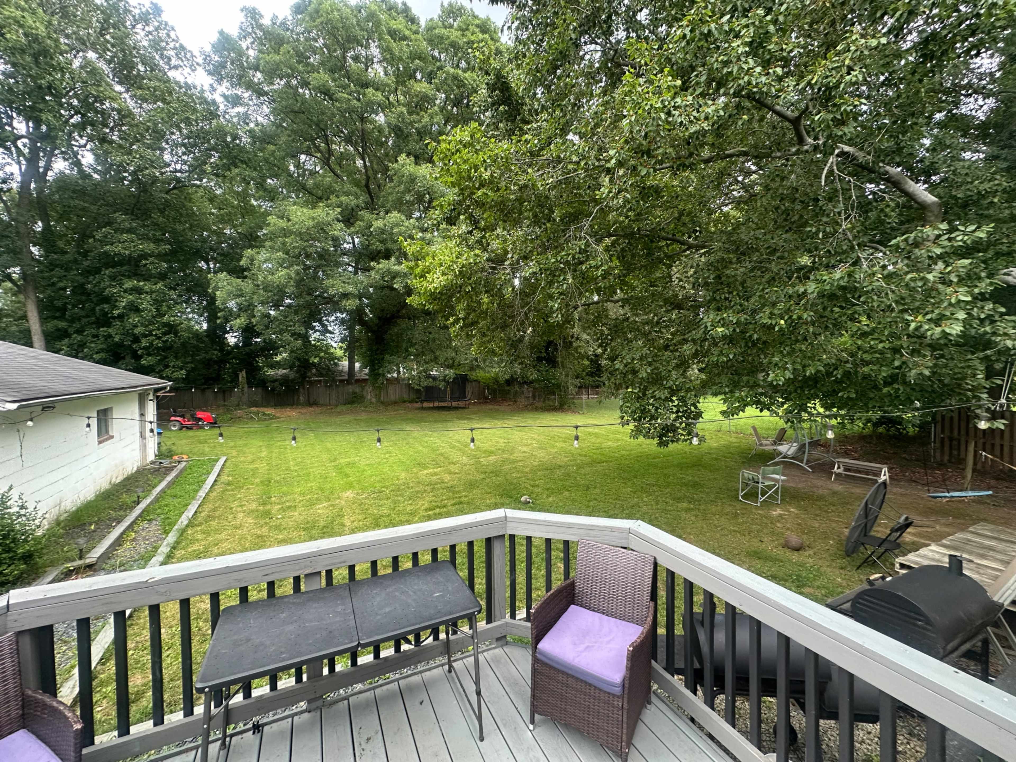 The image shows a backyard view from a deck with a table and chairs, featuring a grassy lawn, trees, and some outdoor furniture.