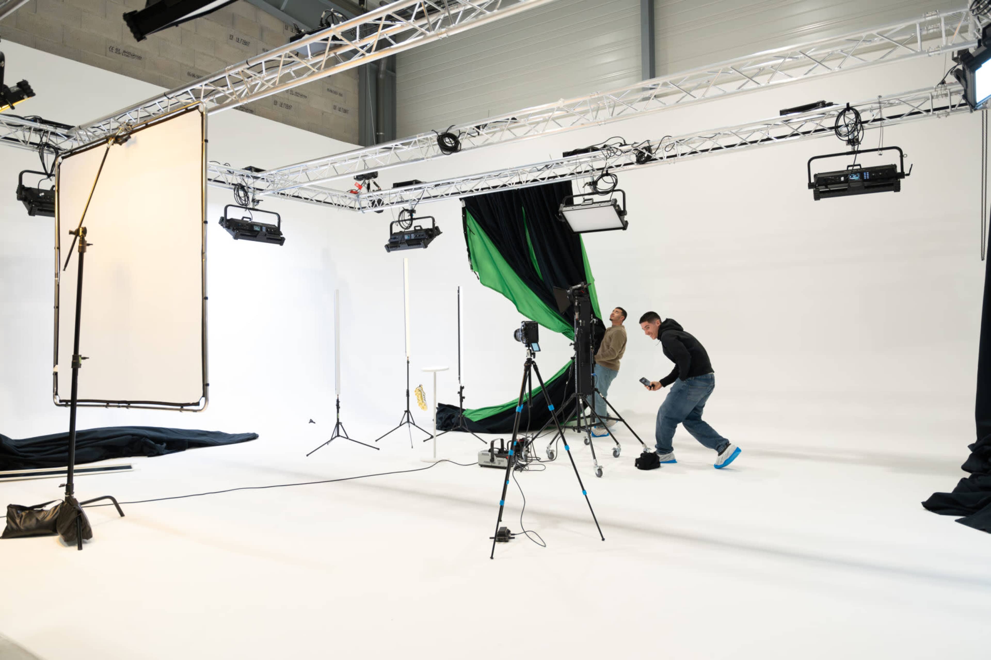A photography studio with a white background is set up with lighting equipment, a green backdrop, and two individuals adjusting the scene.