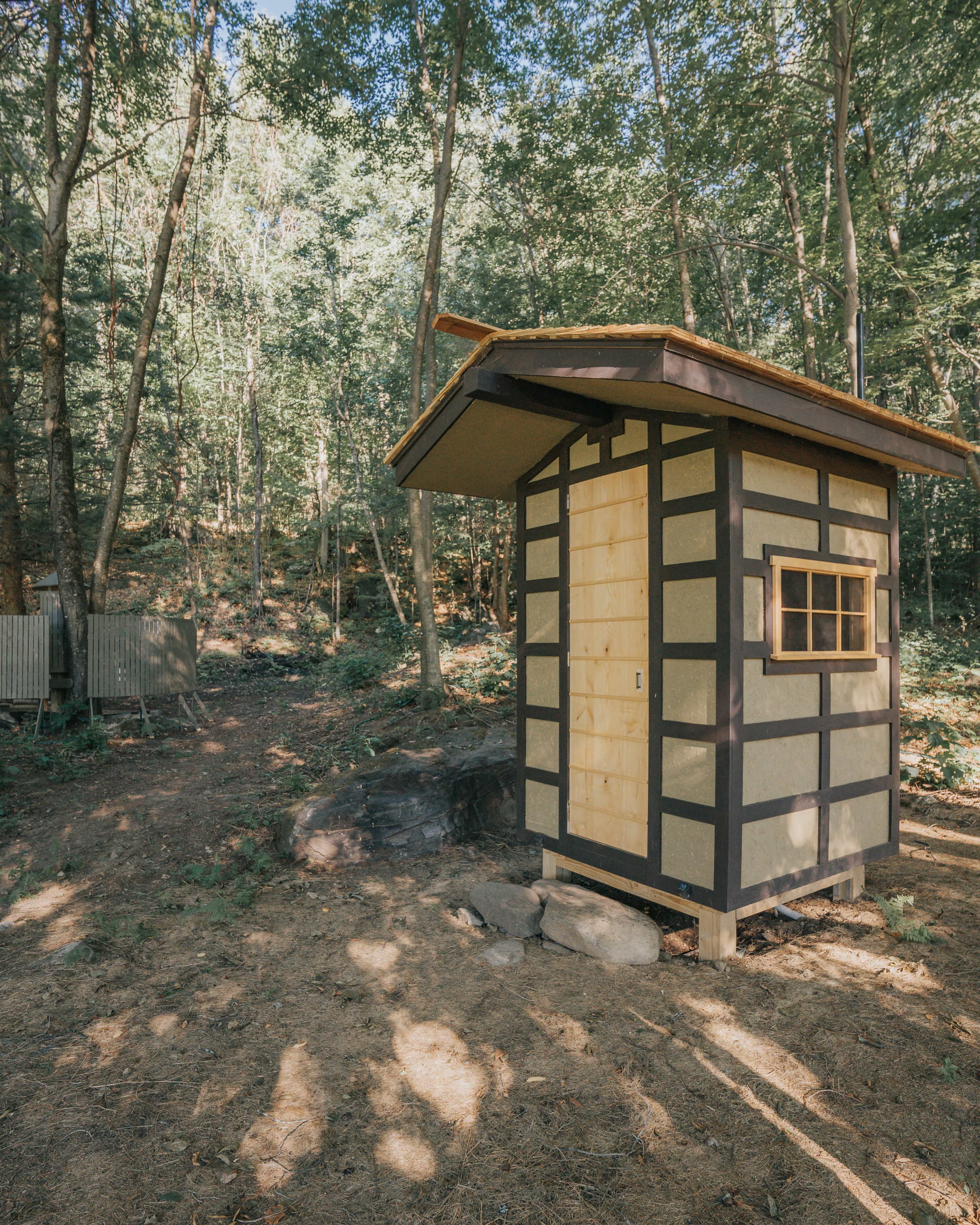 A small, wooden cabin with a slanted roof and a single window is surrounded by trees in a forested area.