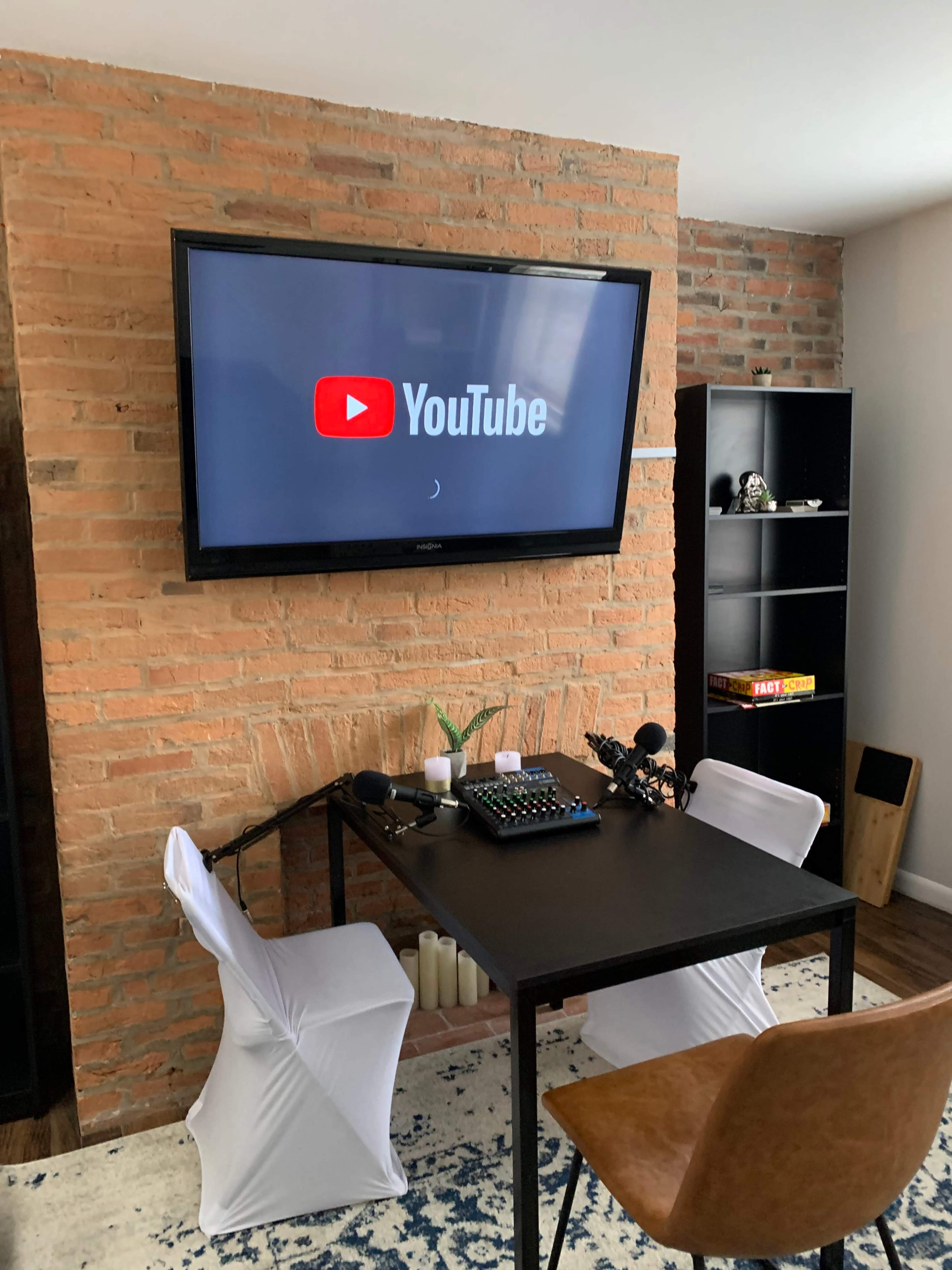 A black table with microphones and a control panel is positioned in front of a television displaying the YouTube logo against a brick wall.
