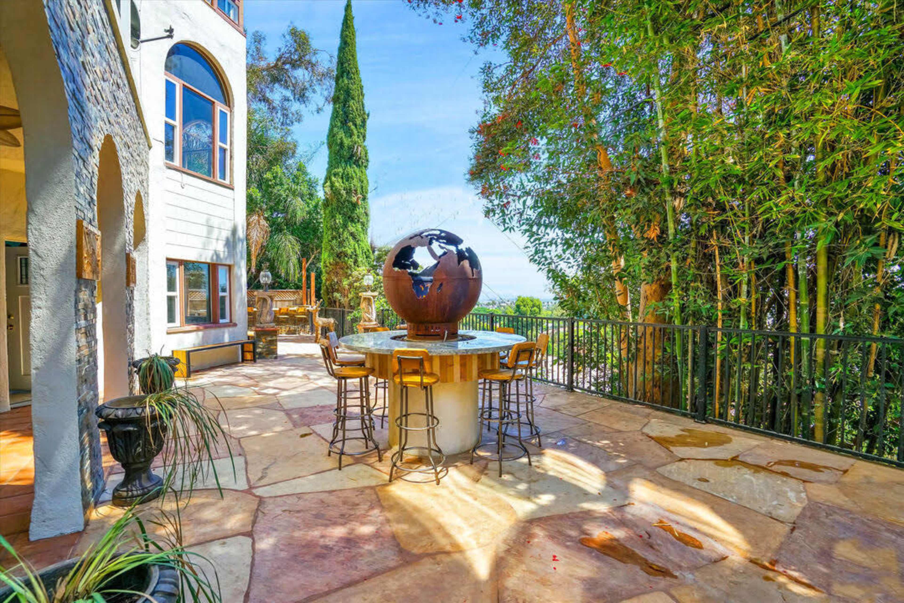 The image shows a spacious outdoor terrace with a round table featuring a globe centerpiece, surrounded by bar stools and framed by lush greenery and a dramatic view.