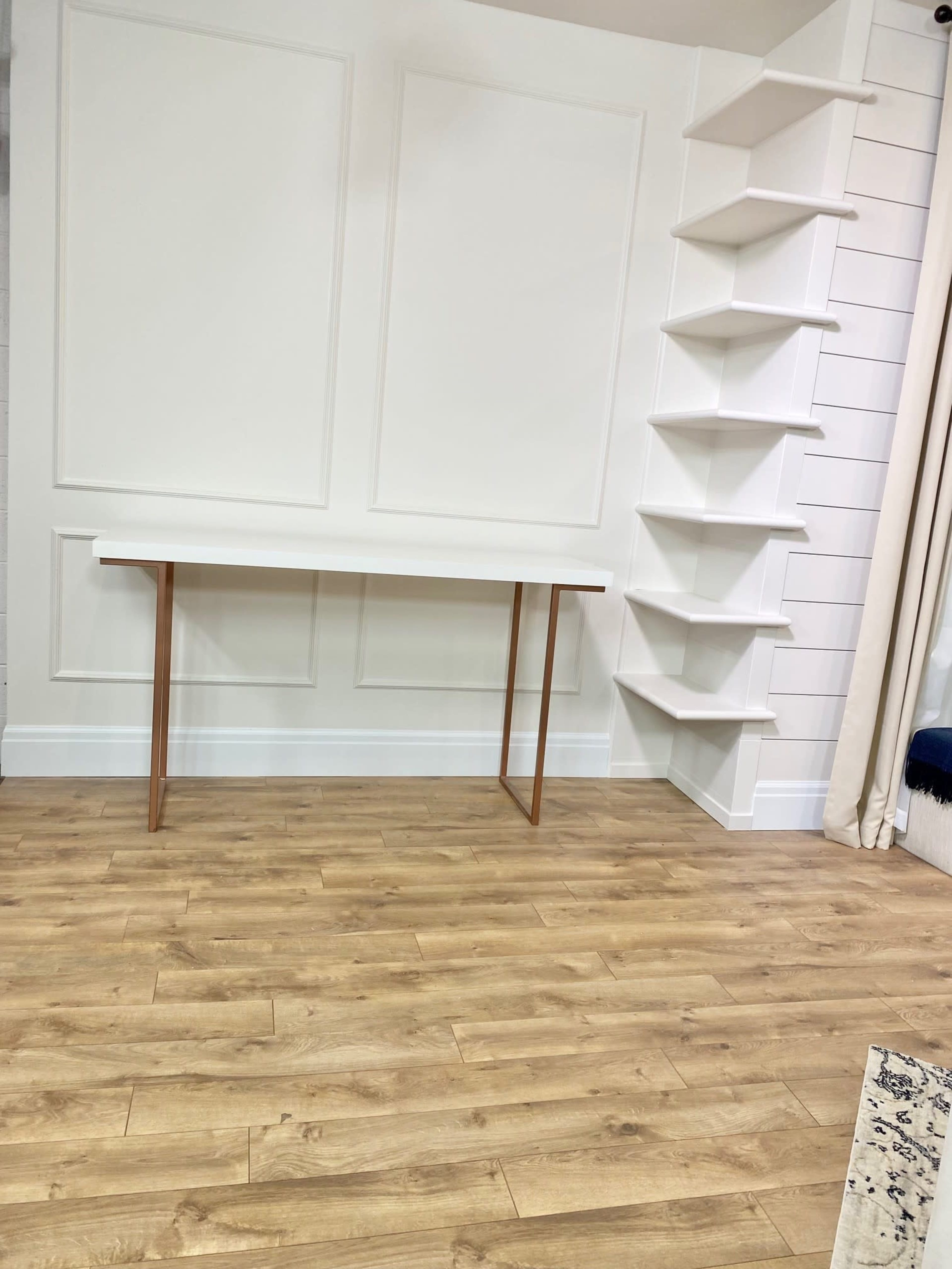 A minimalist room featuring a white table with metal legs and a set of wall-mounted shelves against a paneled wall, with wooden flooring.