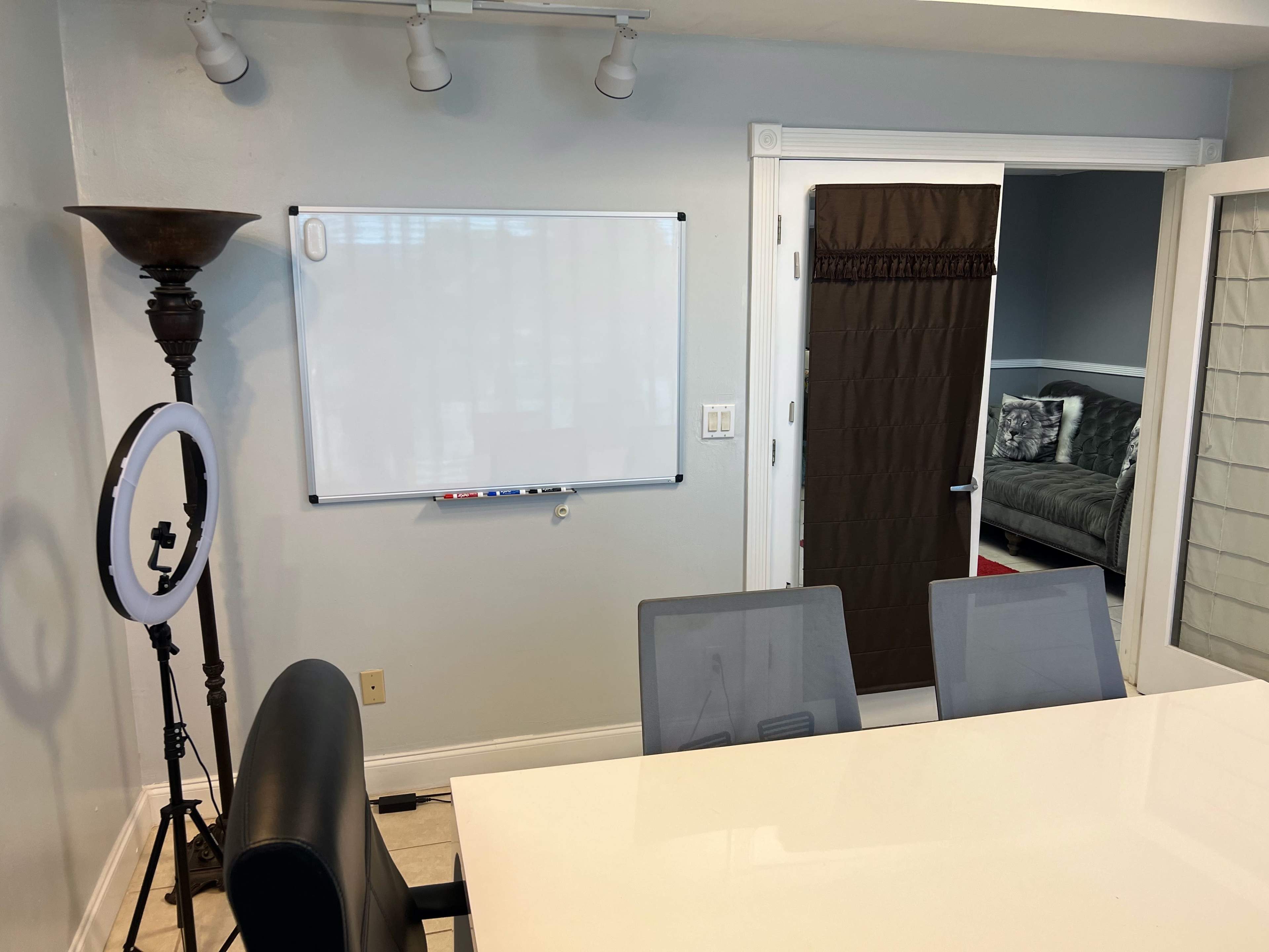 The image shows a meeting room with a whiteboard on the wall, a large table with chairs, and a ring light placed beside a floor lamp.