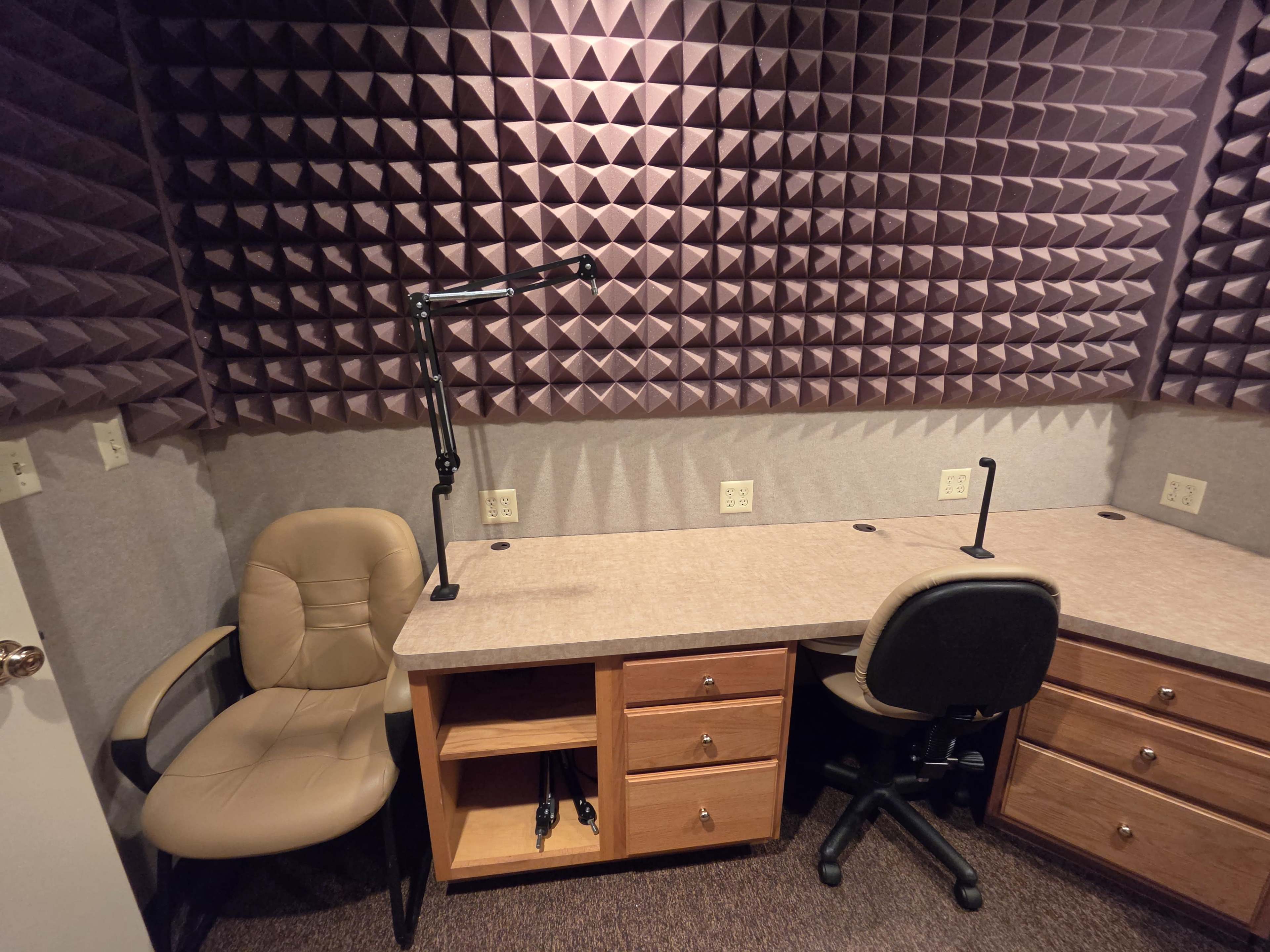 The image shows a small recording studio with soundproof foam panels on the walls, two desks, and two office chairs.