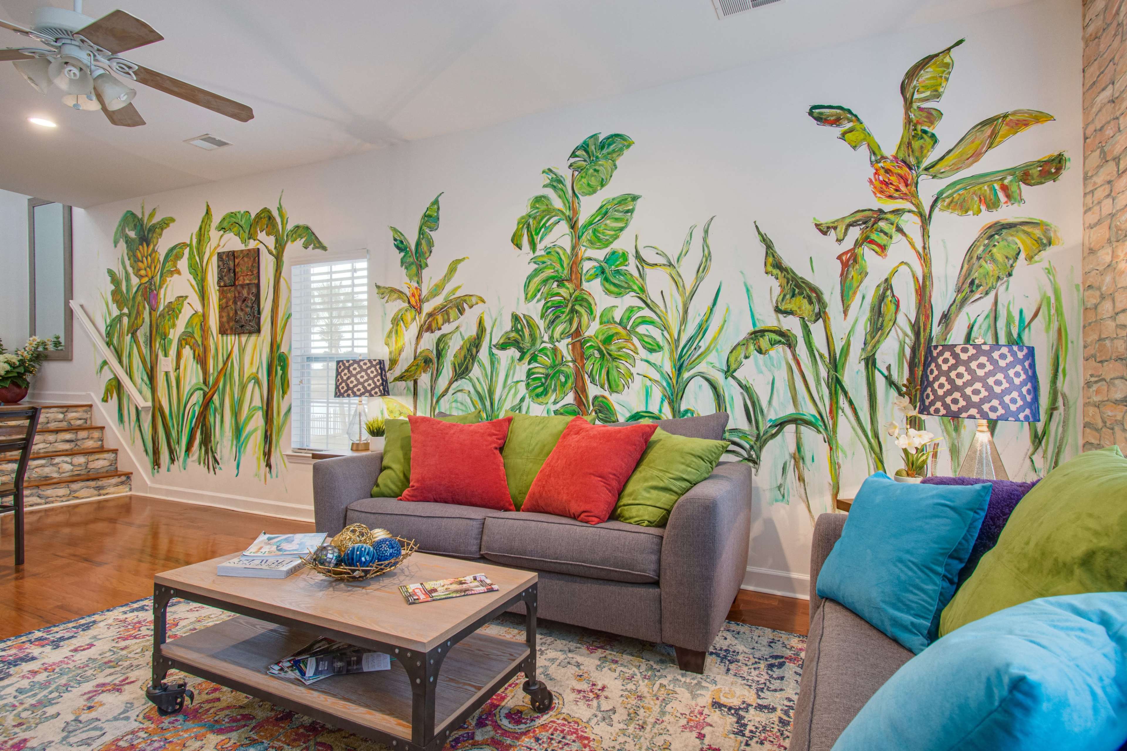 A living room features a gray sofa and a blue sofa, with a wooden coffee table and vibrant plant-themed wall murals.