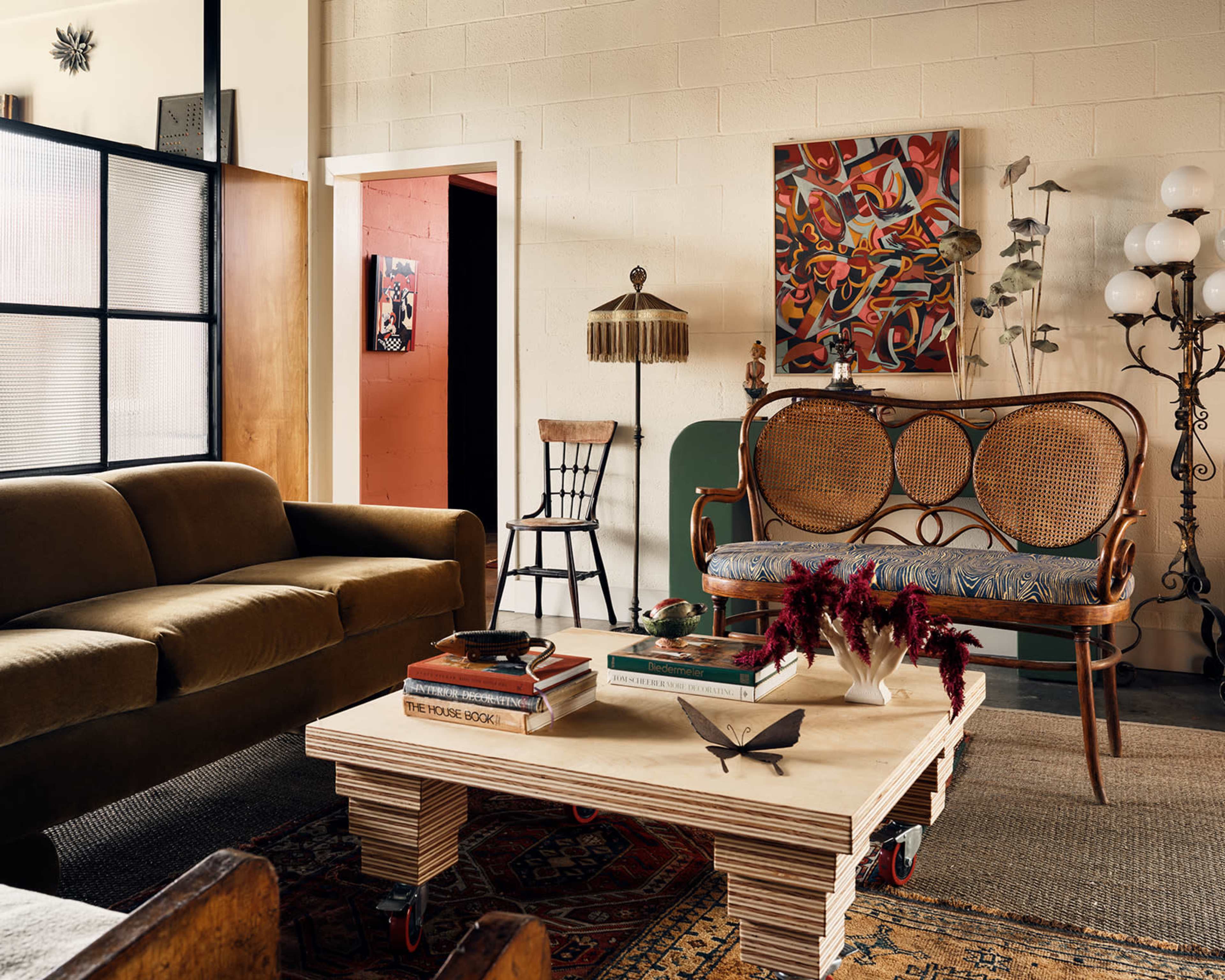 The image shows a cozy living room with a brown sofa, a vintage wooden chair, a coffee table on wheels, and various artworks and plants decorating the space.