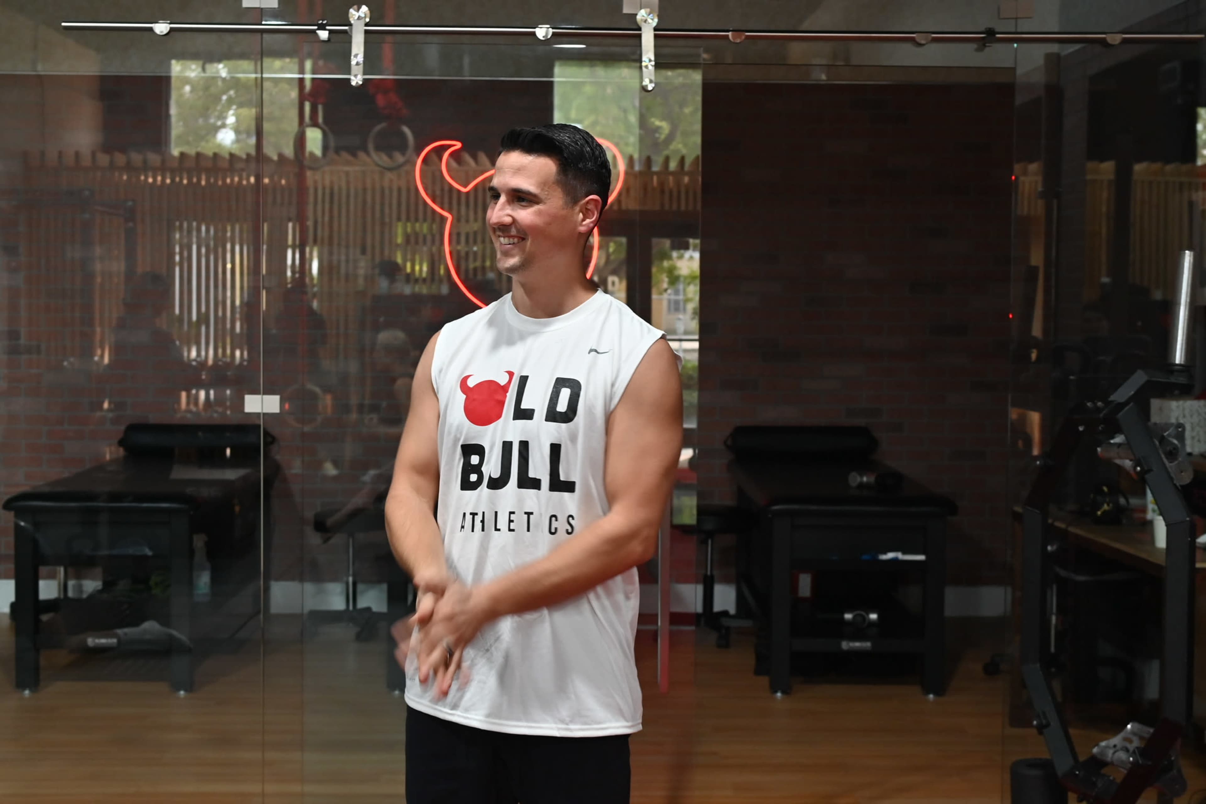 A man wearing a sleeveless shirt stands in a gym, smiling and clapping his hands, with a neon bull logo illuminated in the background.