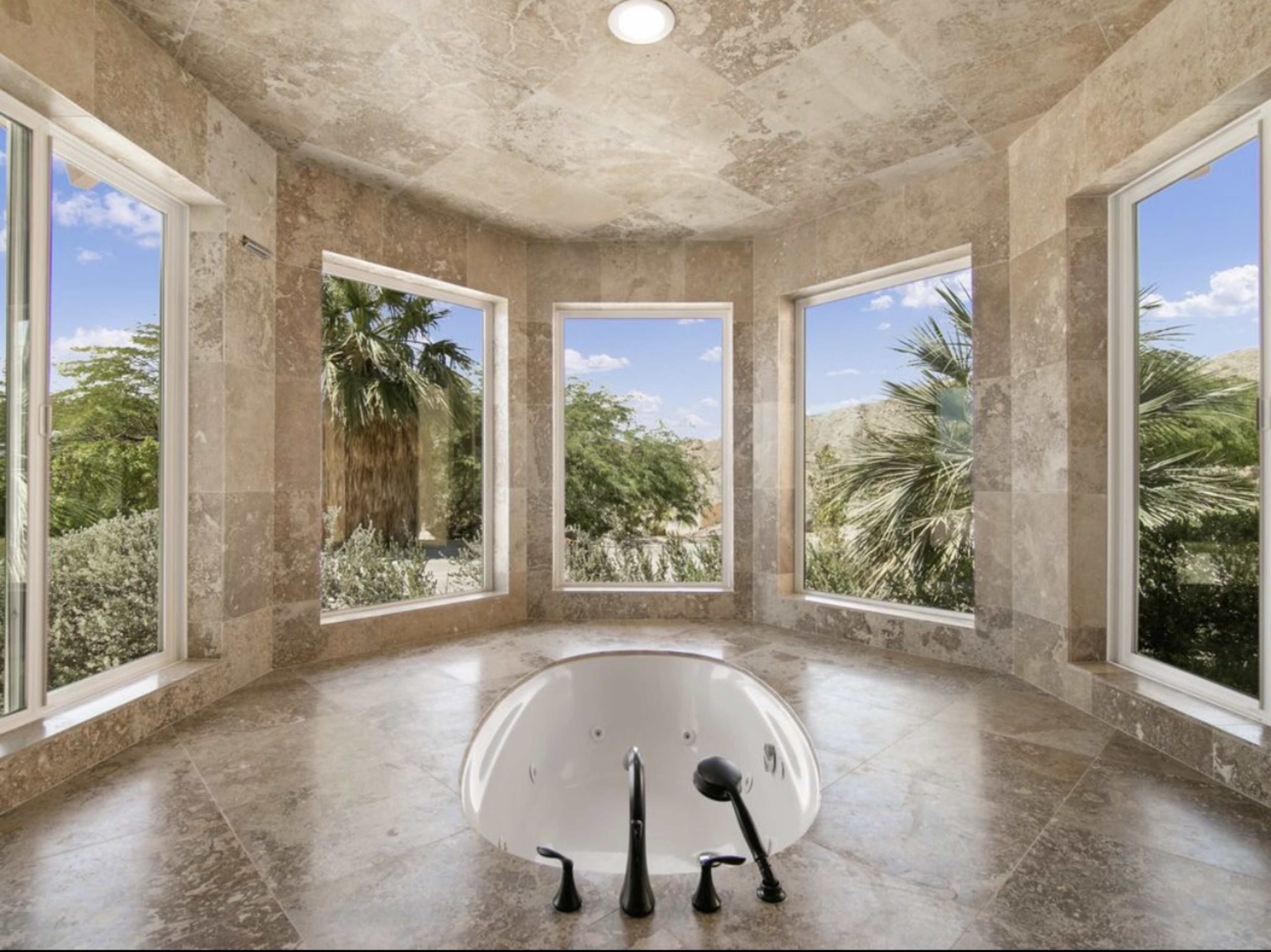 A soaking tub is positioned in a corner of a spacious bathroom featuring large windows that provide a view of surrounding palm trees and greenery.
