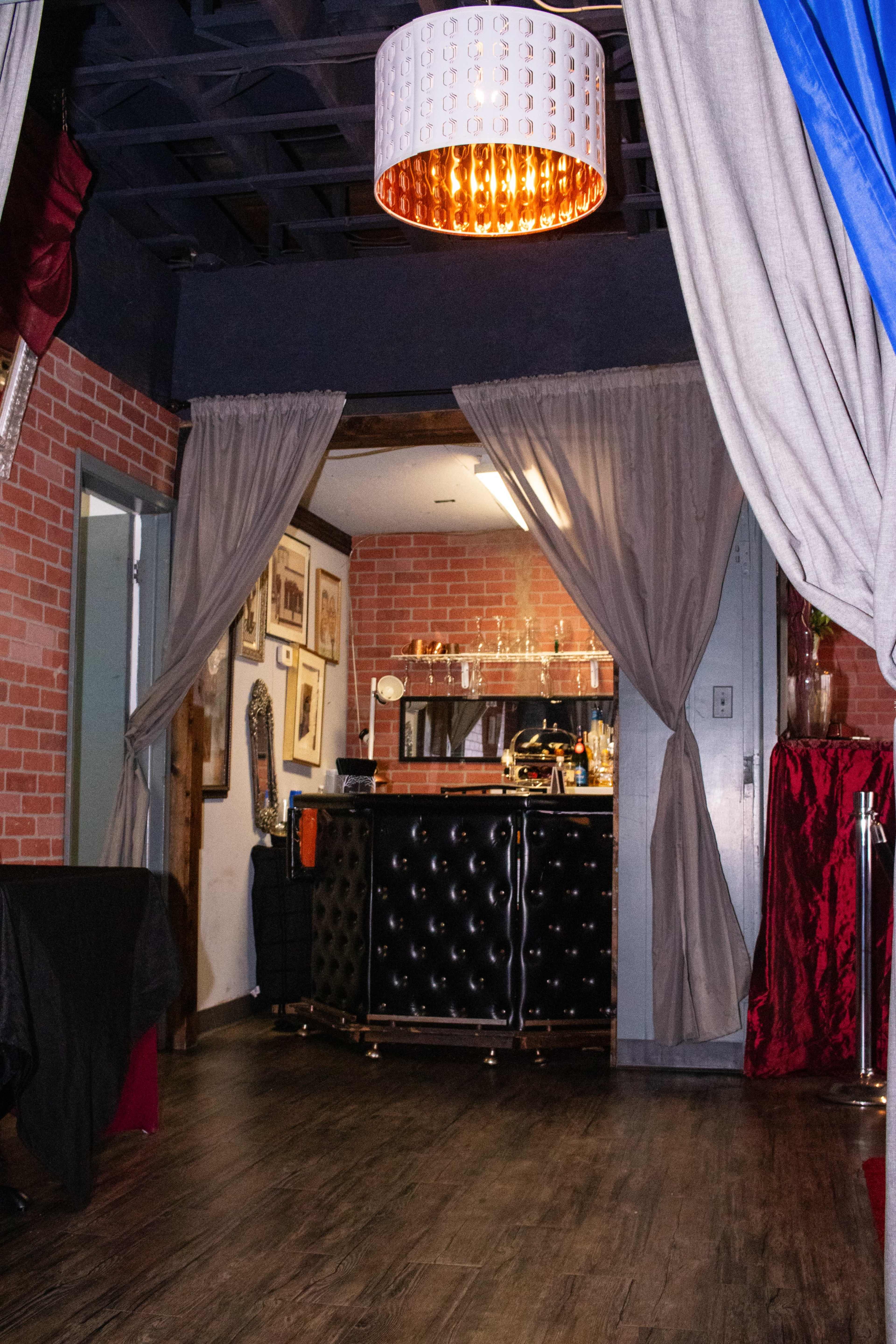 The image shows a stylish interior featuring a bar area with a black tufted counter, framed artwork on the walls, and two curtains framing the entrance.