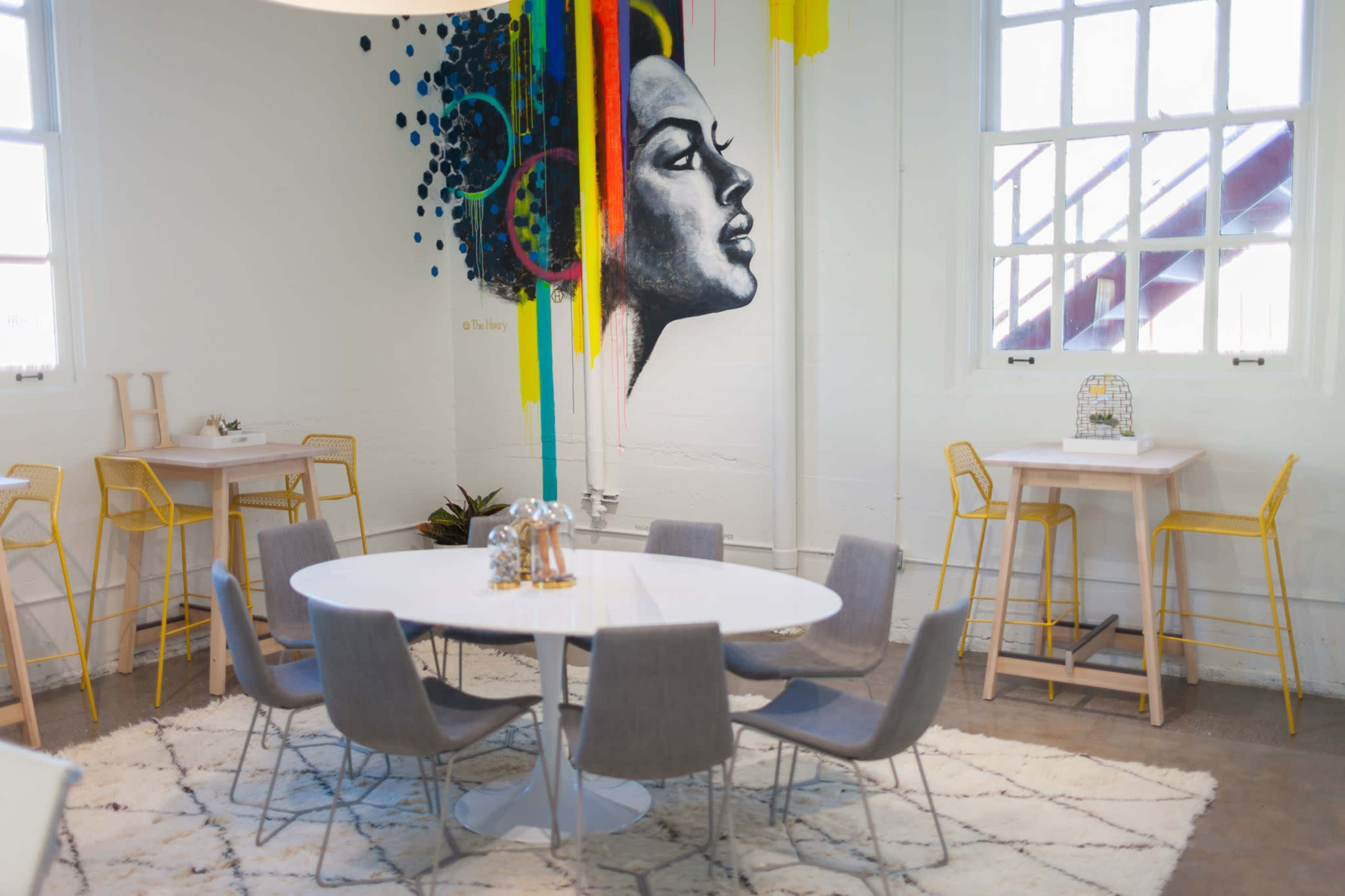A modern workspace with a round white table surrounded by six gray chairs, yellow bar stools, and a colorful mural depicting a woman's profile on the wall.