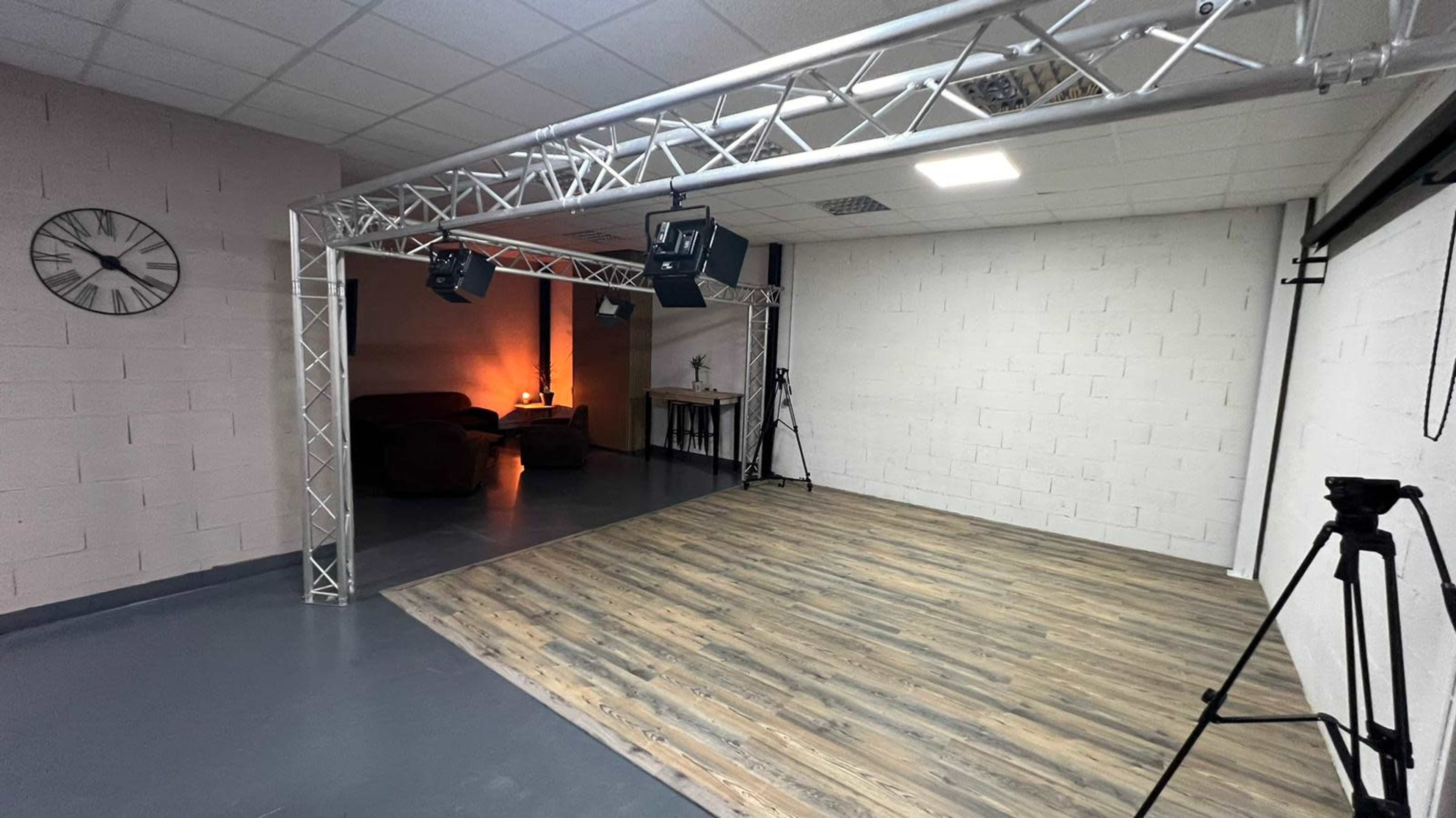 An empty room with wooden flooring, a wall-mounted clock, and lighting equipment set up for a studio or performance space.