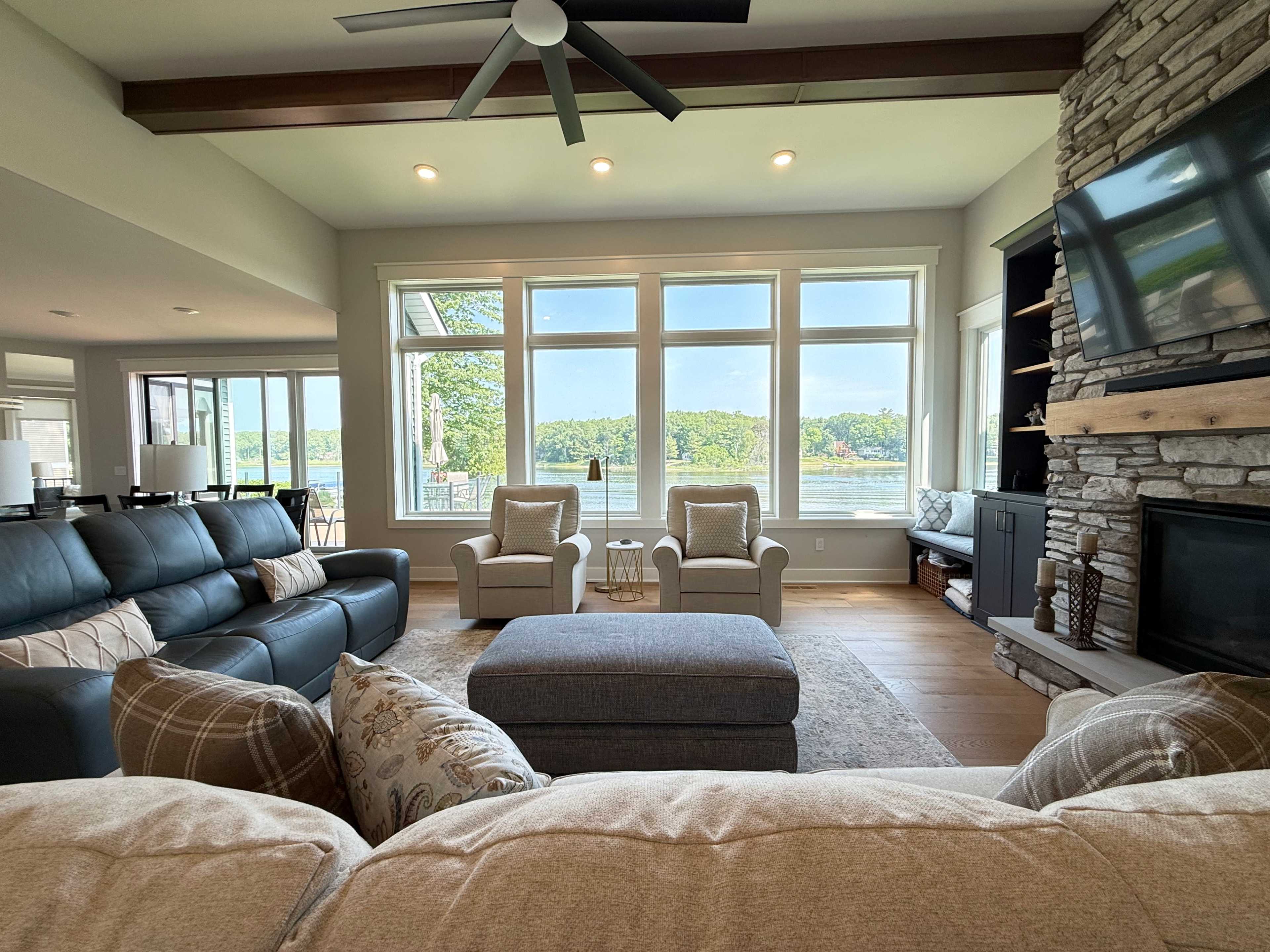 The image shows a modern living room with a view of a lake, featuring comfortable seating, a stone fireplace, and large windows allowing natural light.