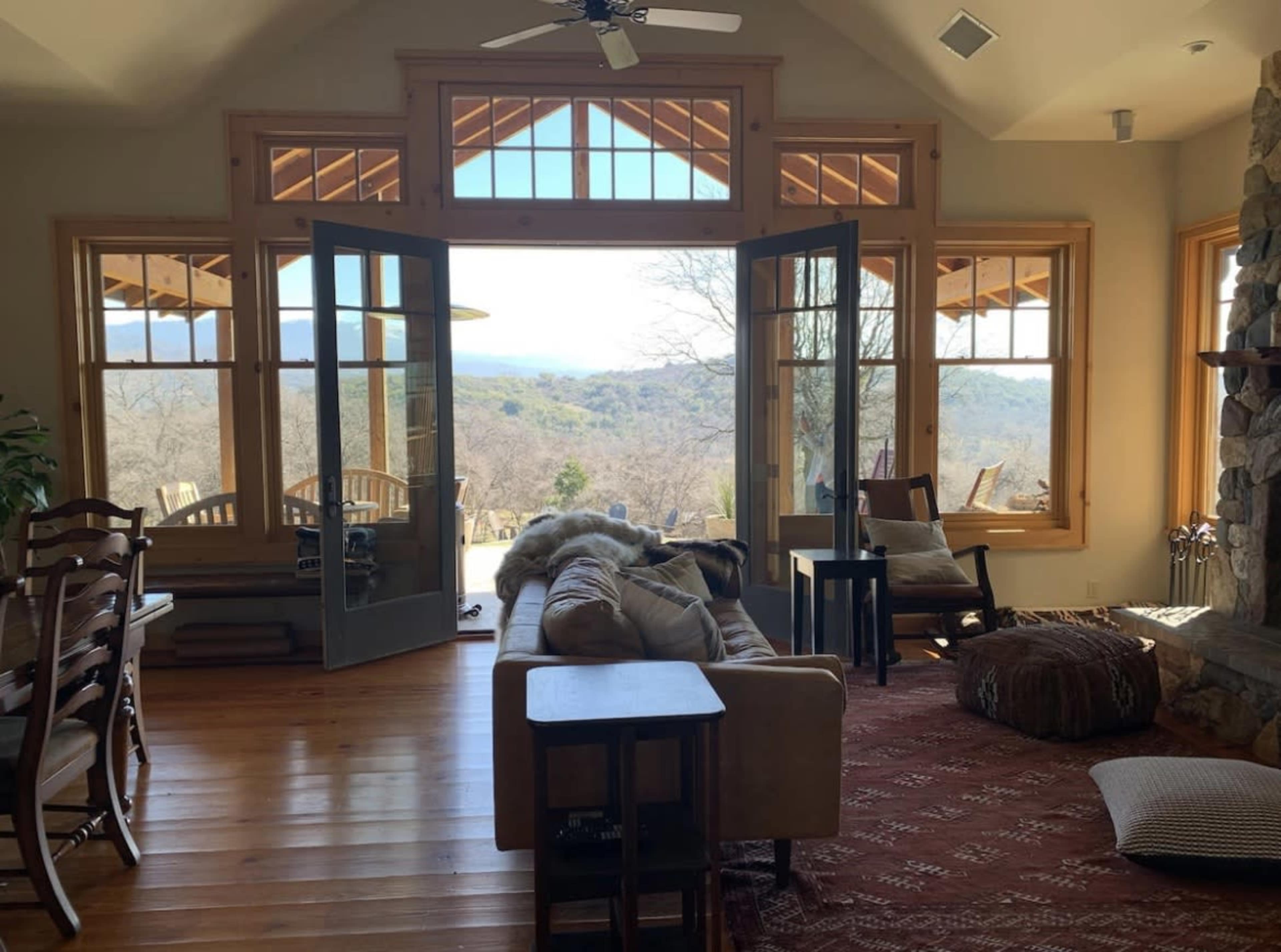A spacious living room features large windows that open to a scenic view, with a sofa, dining table, and various seating arrangements.