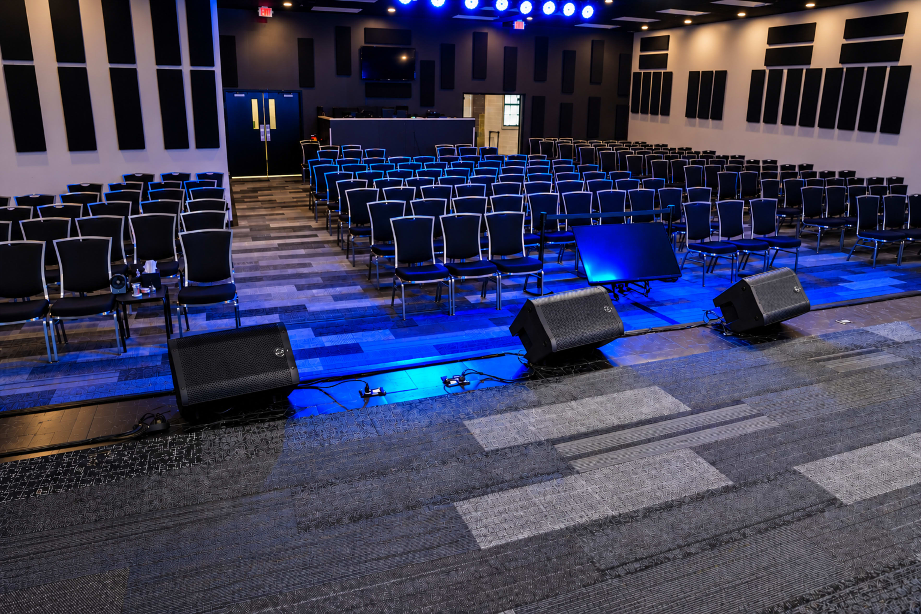 The image shows a spacious auditorium with rows of empty chairs facing a stage, equipped with two speakers and blue lighting.
