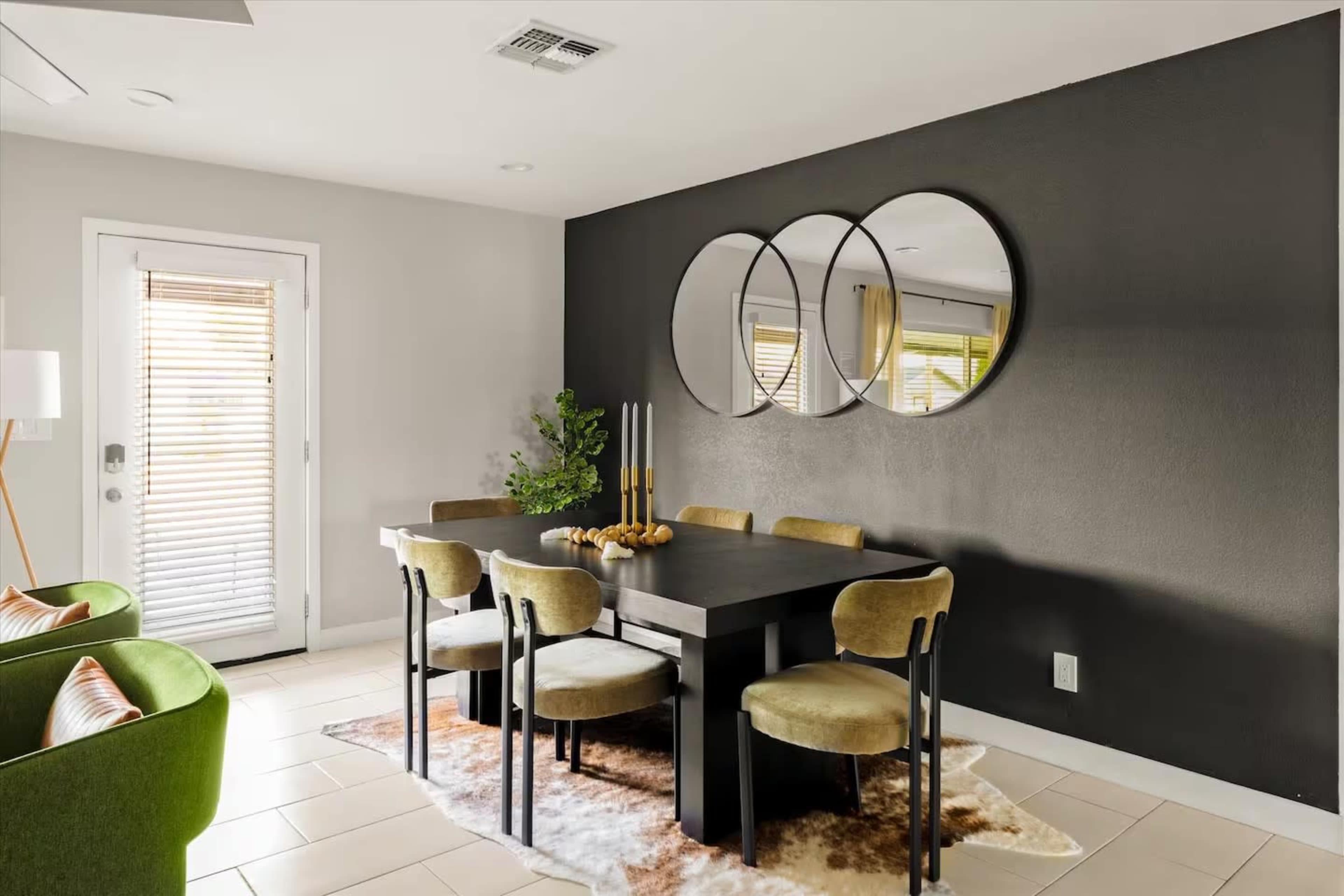 The image shows a dining area featuring a dark wall, a rectangular table surrounded by chairs, and three circular mirrors above the table.