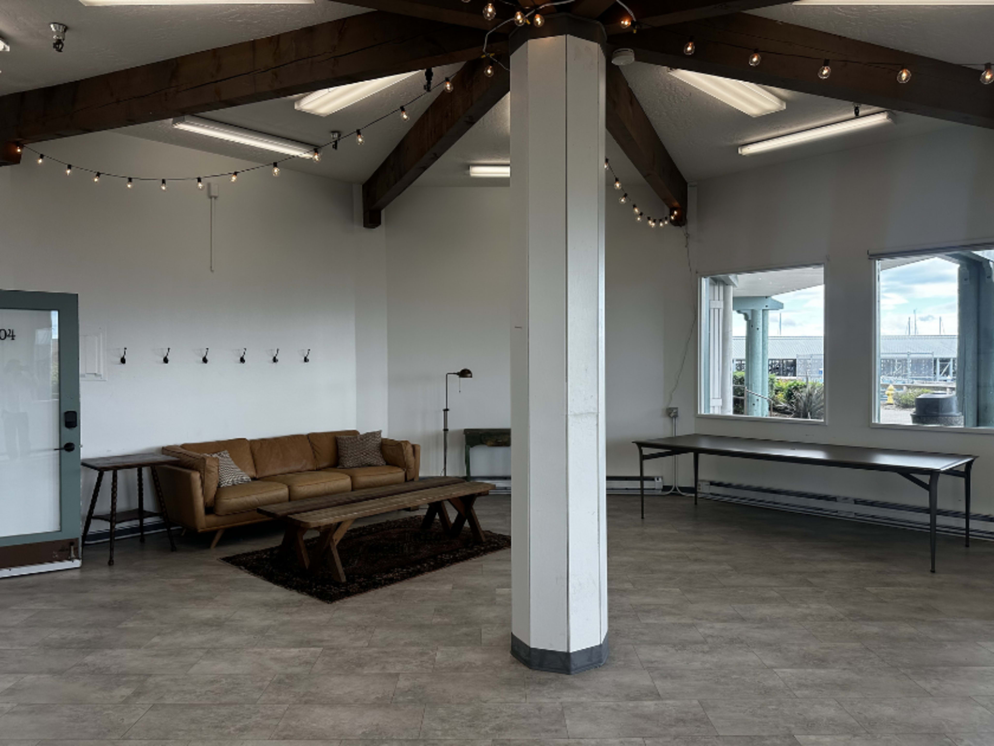 The image shows a spacious interior with a tan couch, a wooden coffee table on a dark rug, a table against the wall, and large windows letting in natural light.