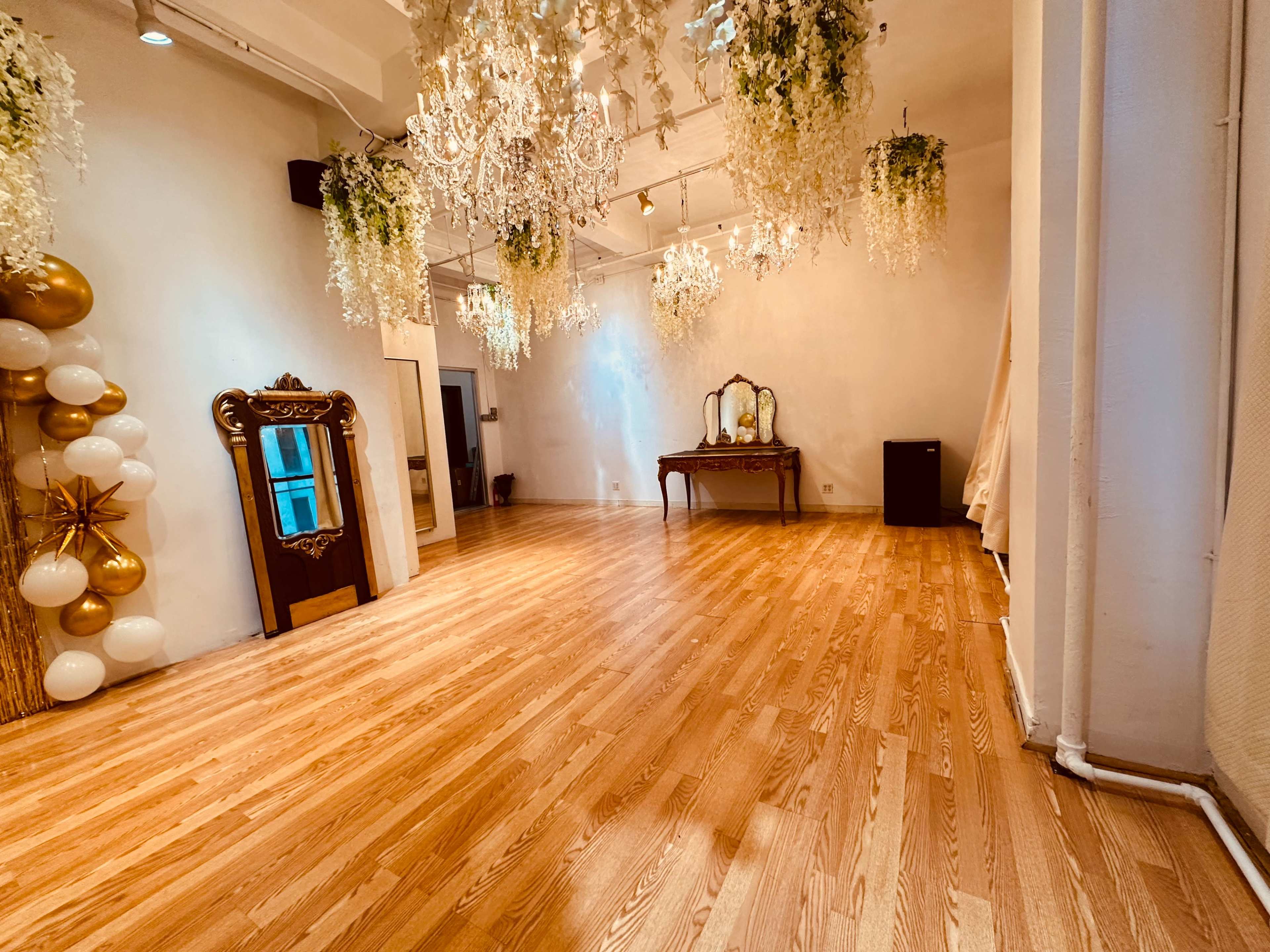 The image shows a spacious, well-lit room with wooden floors, decorated with chandeliers and greenery hanging from the ceiling, featuring a large mirror and a vintage-style vanity table against the wall.